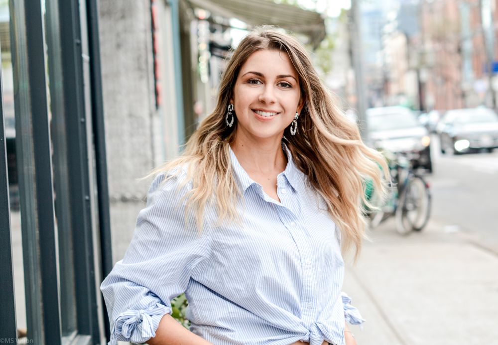 Woman in light blue button down shirt posing casually outdoors on urban street.