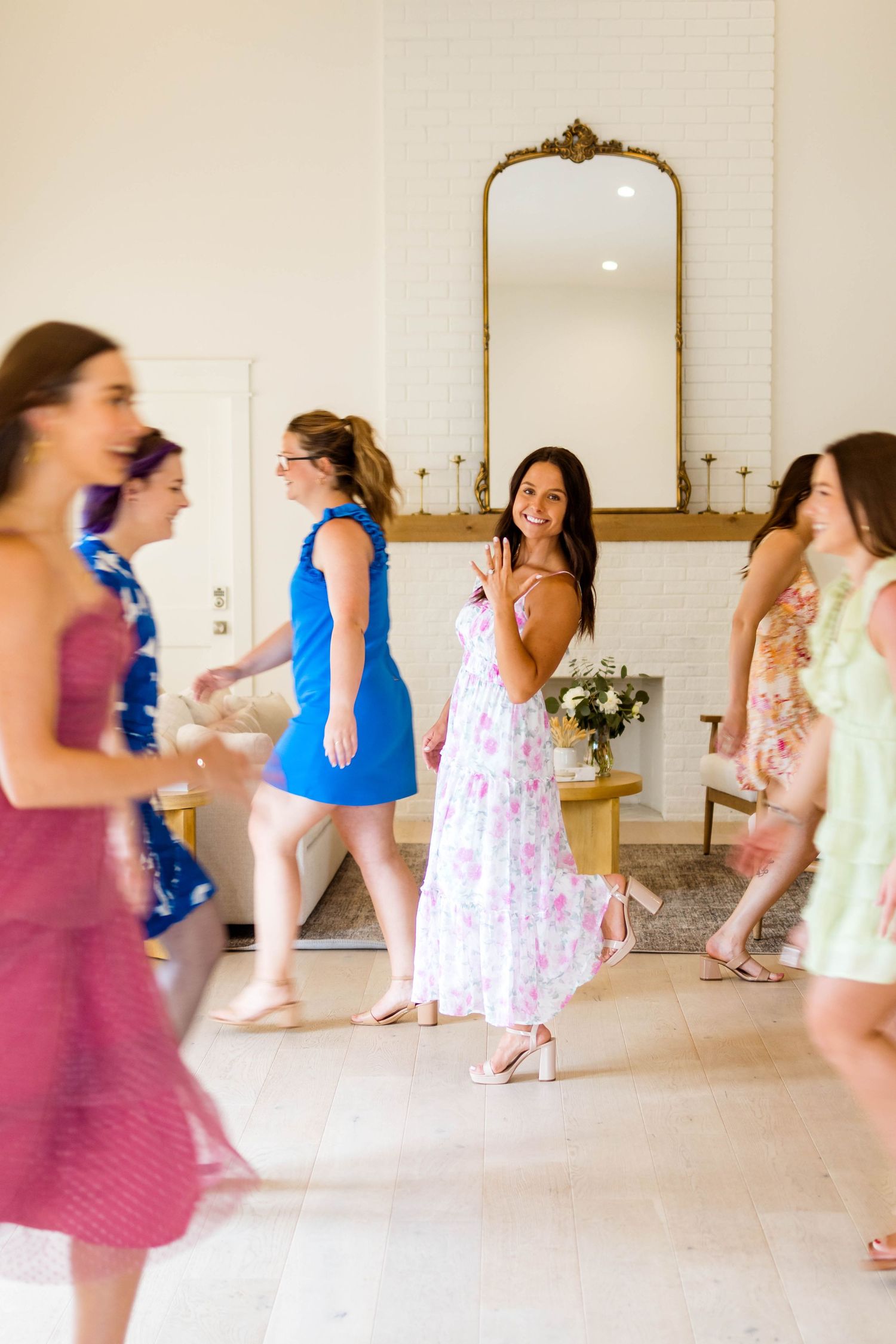 A group of women in colorful summer dresses dancing together in a bright white room with ornate gold mirrors.