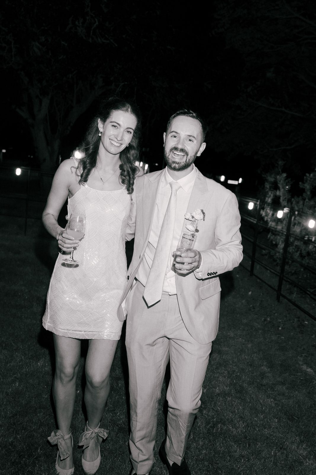A black and white photo of two people at an outdoor event, with one in a white suit and one in a short dress.