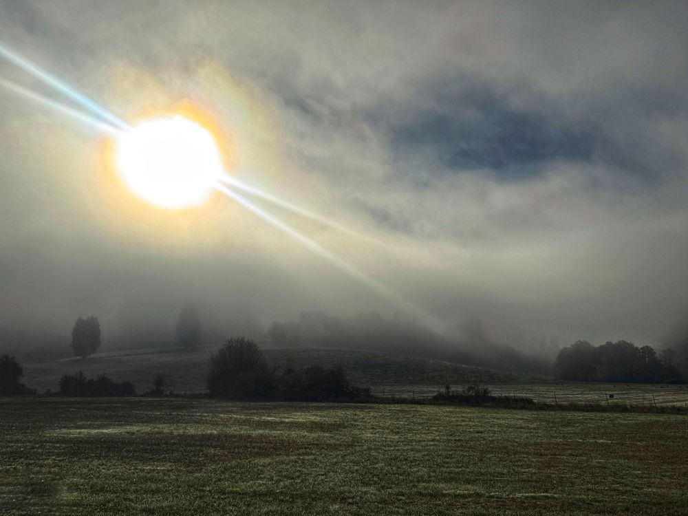A dramatic panoramic view of a misty field at sunrise with dark clouds and golden sunlight breaking through the atmosphere.