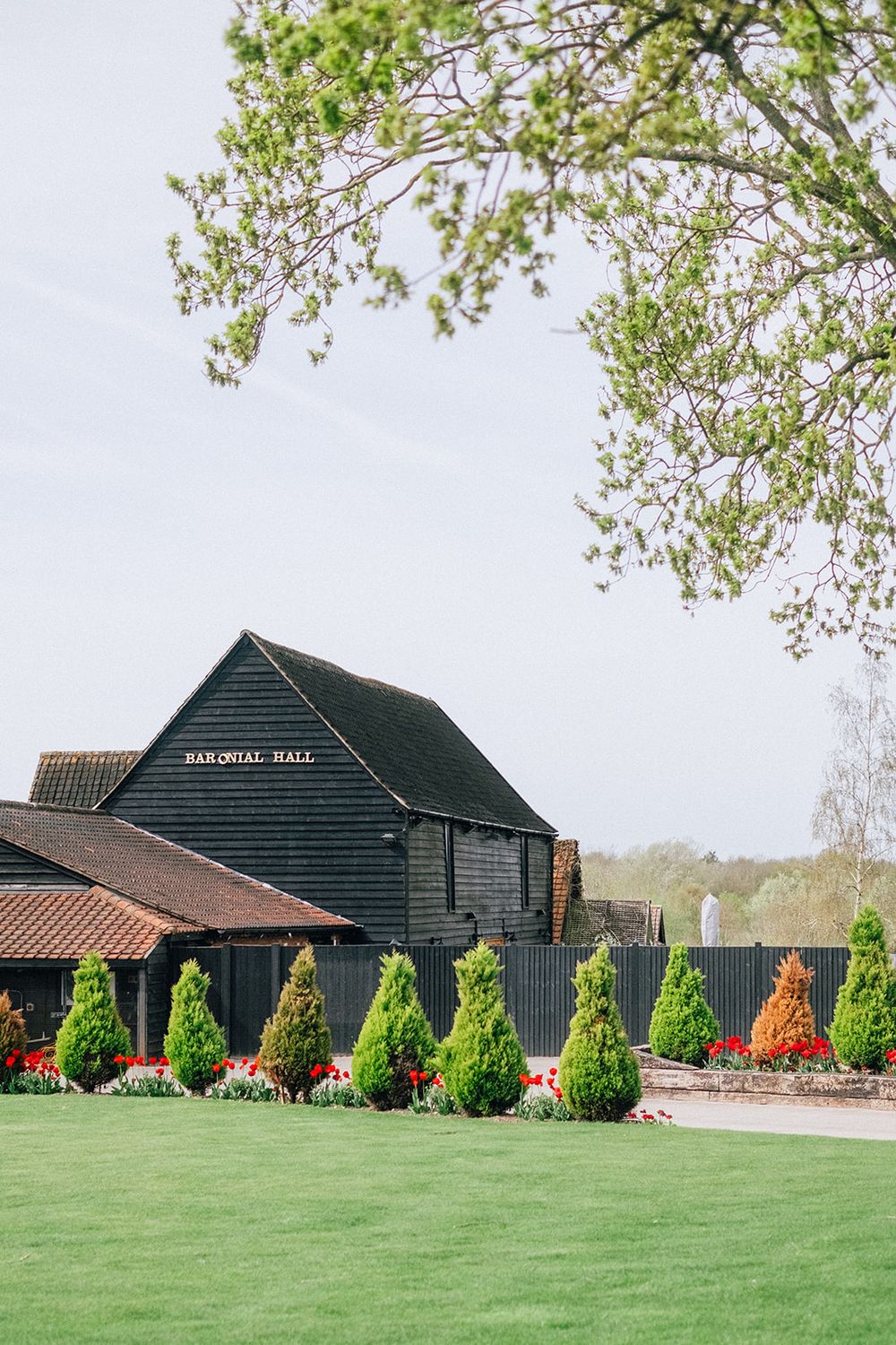 Panoramic view of a rustic black barn building with manicured green lawn and decorative topiary bushes in front.