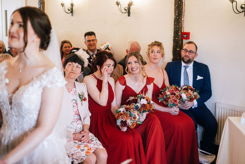 A wedding party in burgundy red dresses poses together holding colorful floral bouquets in an elegant indoor setting.