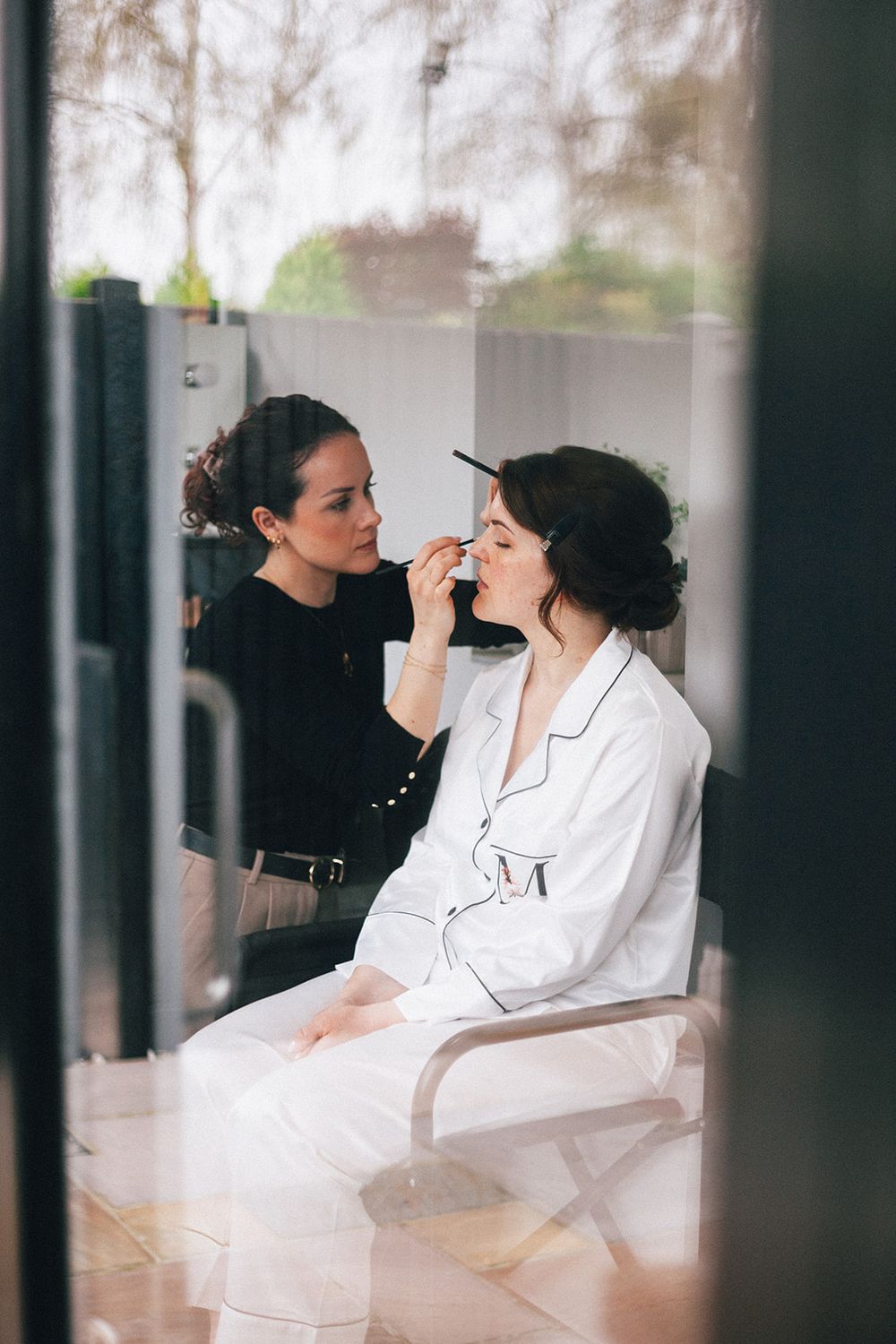 A makeup artist applies eye makeup to someone wearing a white robe during wedding preparations.