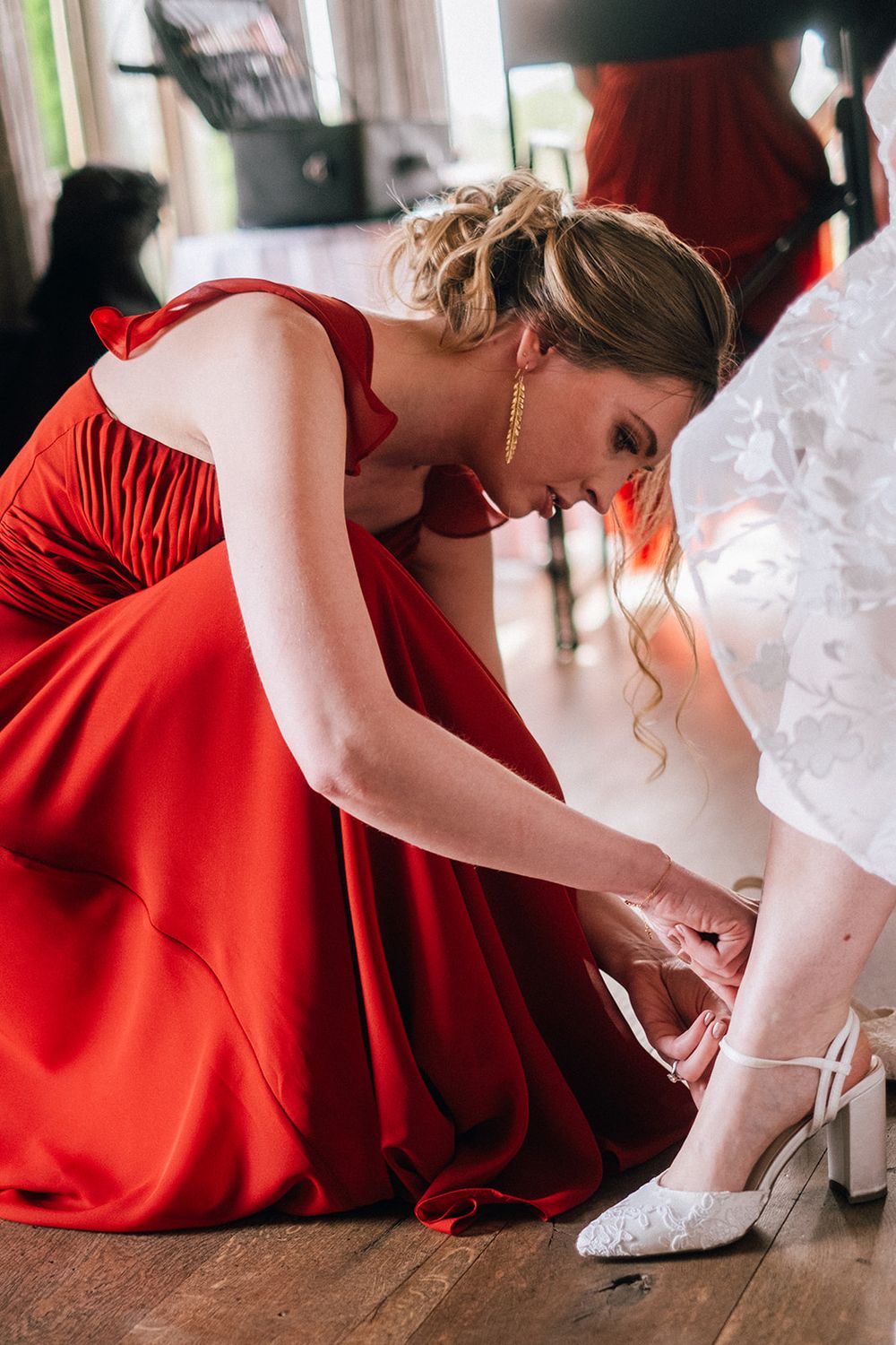 A bridesmaid in a red dress helps adjust a bride's white shoe during wedding preparations.