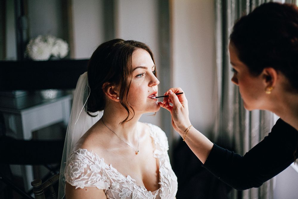 A bride getting her makeup professionally applied in a bright window-lit room while wearing a white lace gown.