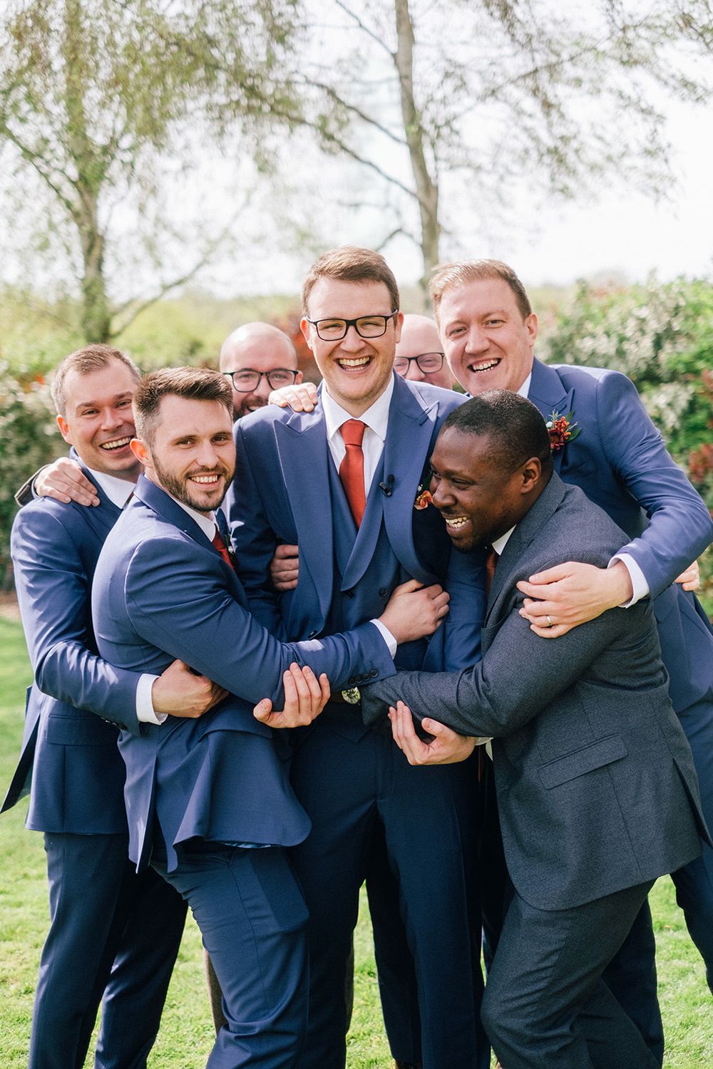 Groomsmen in navy blue suits share a joyful group hug and laugh together outdoors on a sunny day.