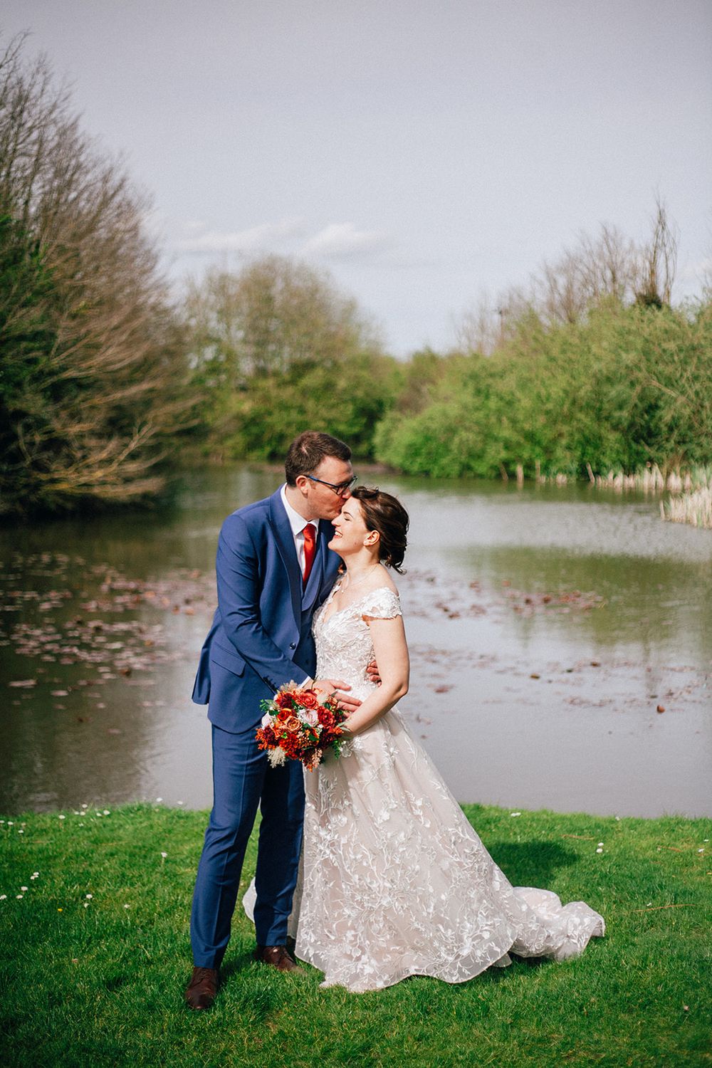 A couple shares a romantic moment in formal attire by a serene lake surrounded by spring greenery.