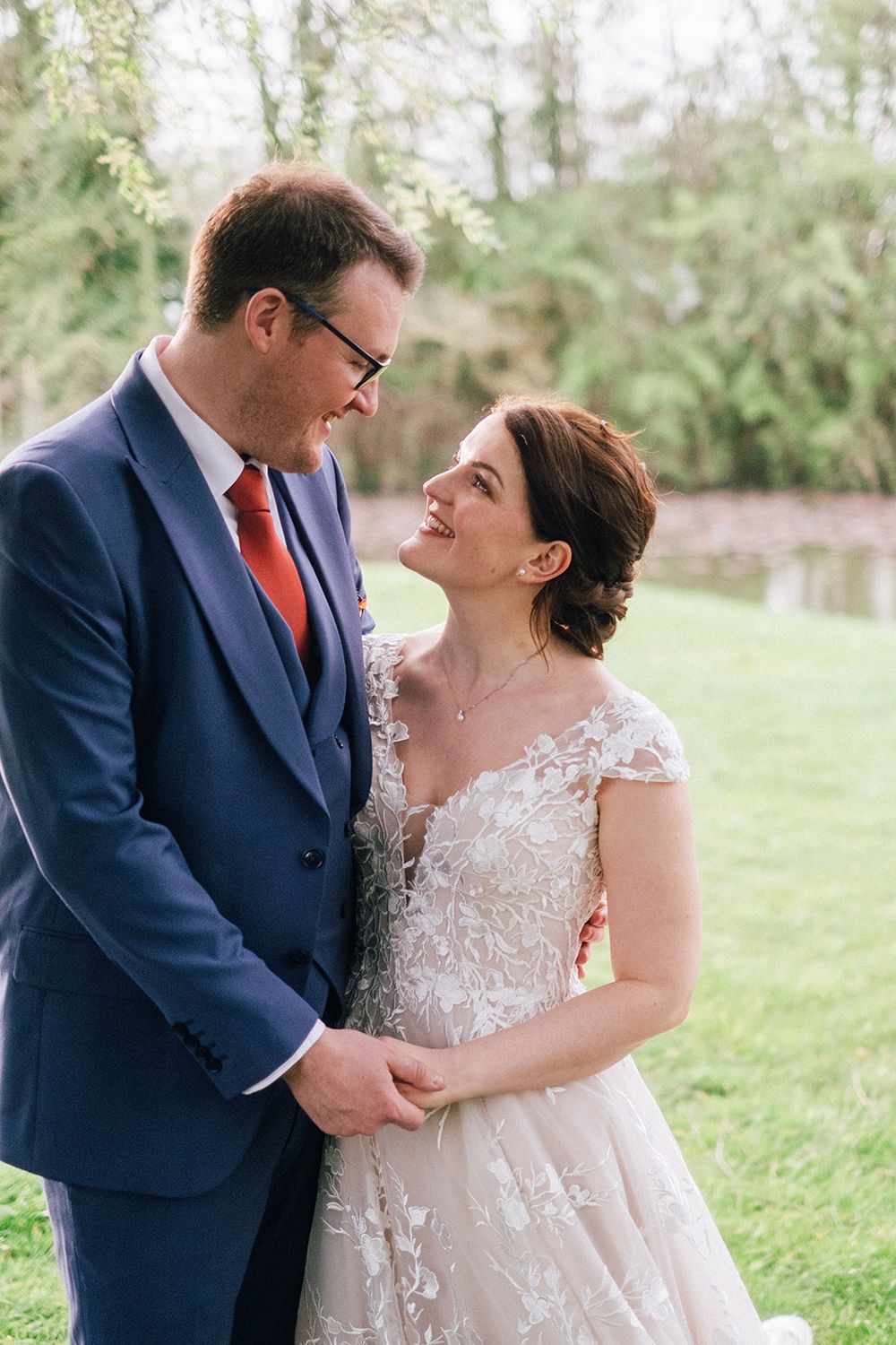 A couple in wedding attire share an intimate moment outdoors, with the bride in lace and groom in navy suit.