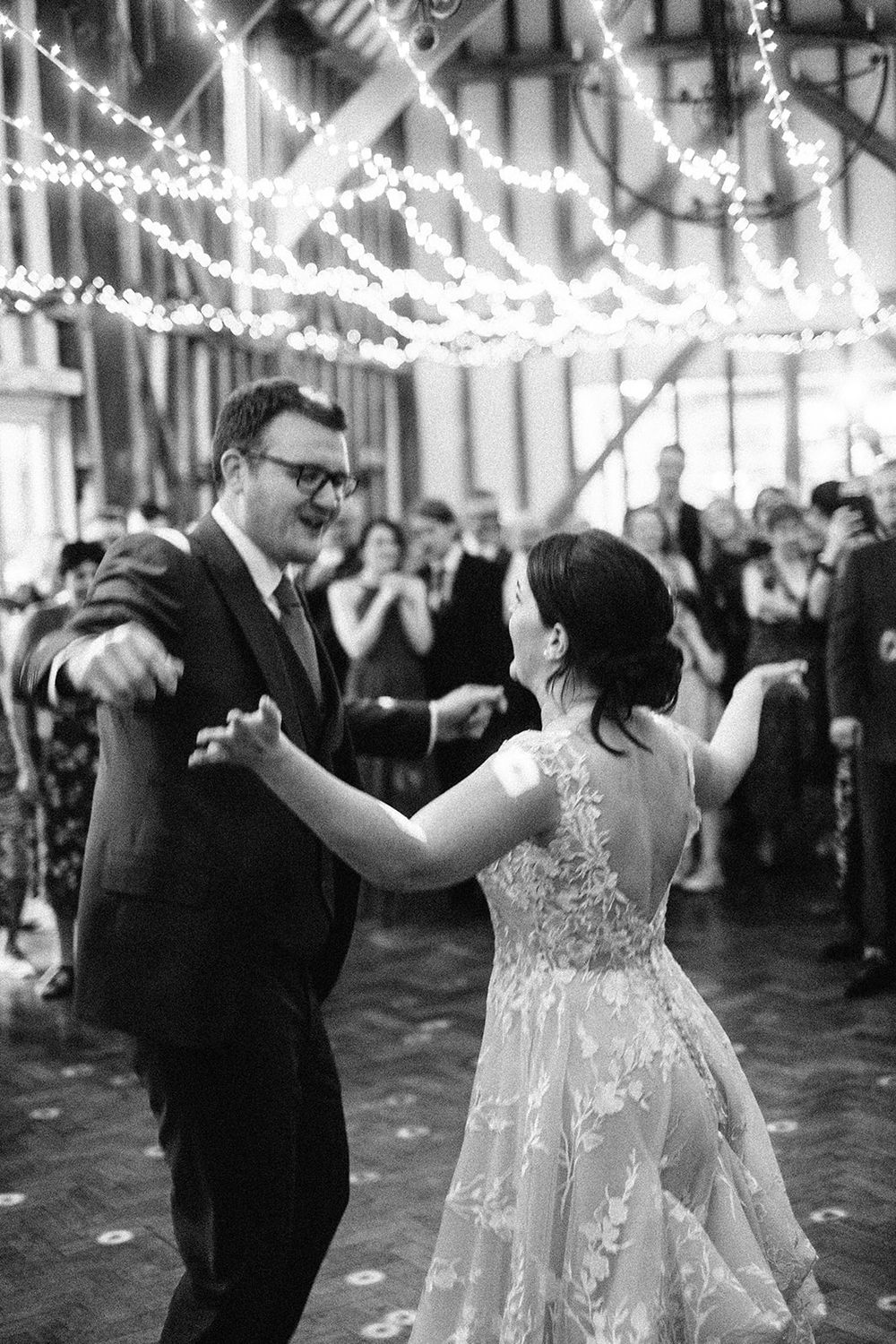 A couple dances together at their wedding reception under twinkling lights, with the bride wearing a lace gown.