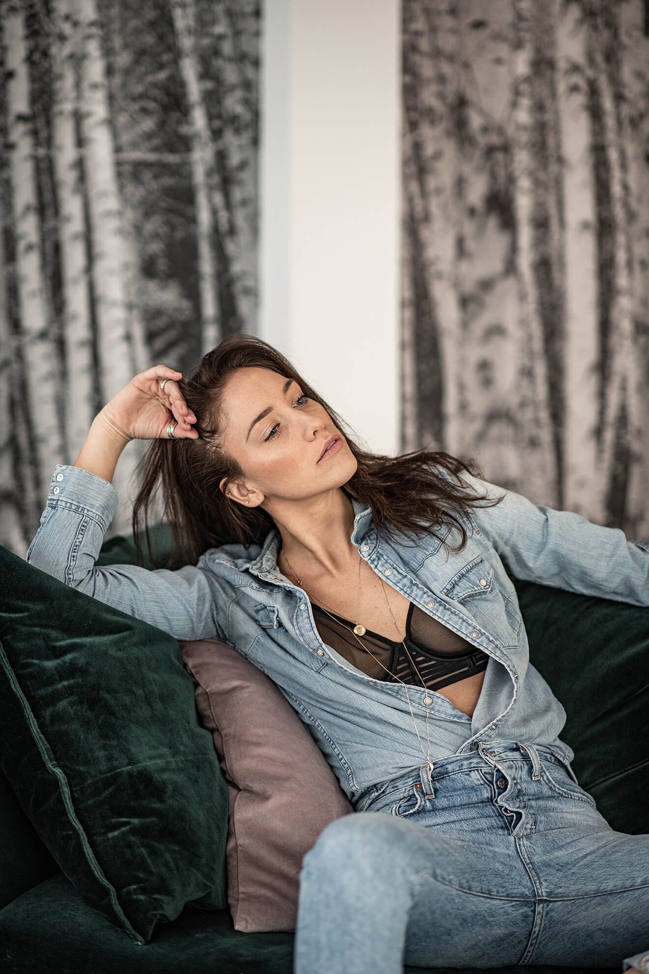 Fashion model relaxes on dark couch wearing casual denim outfit.