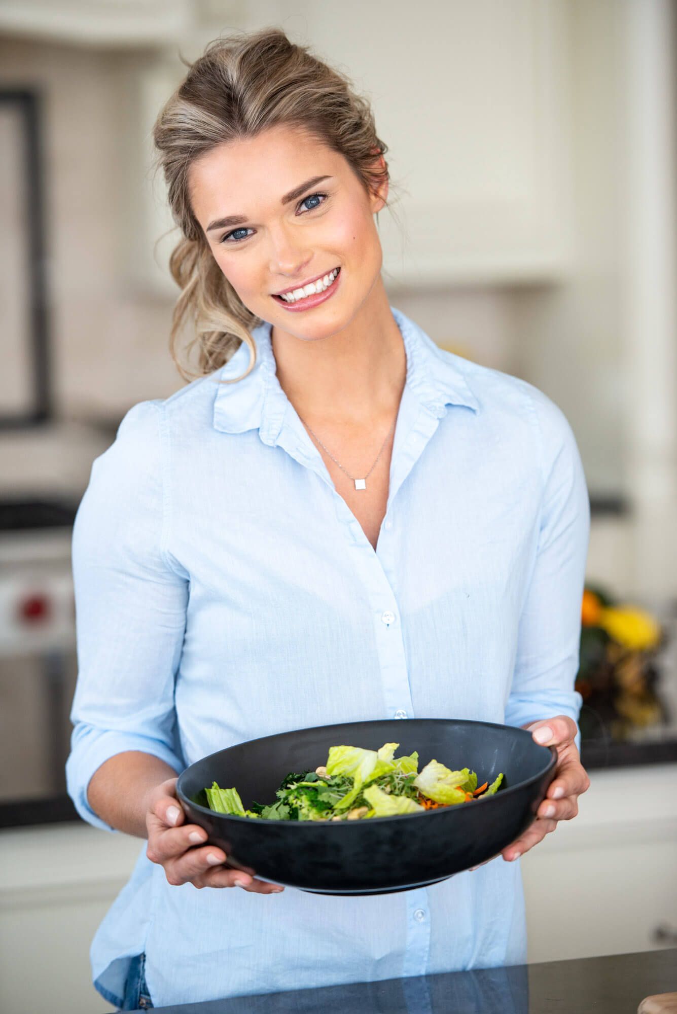 Woman in light blue shirt presenting fresh salad in black bowl in modern kitchen.