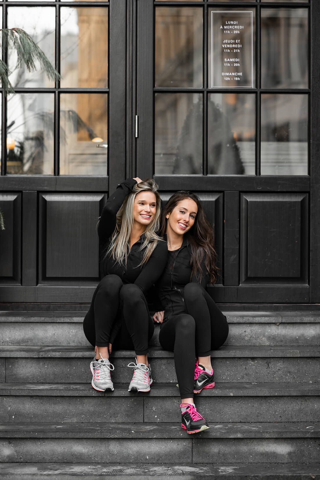 Two friends in black athletic wear pose casually on steps outside storefront windows.