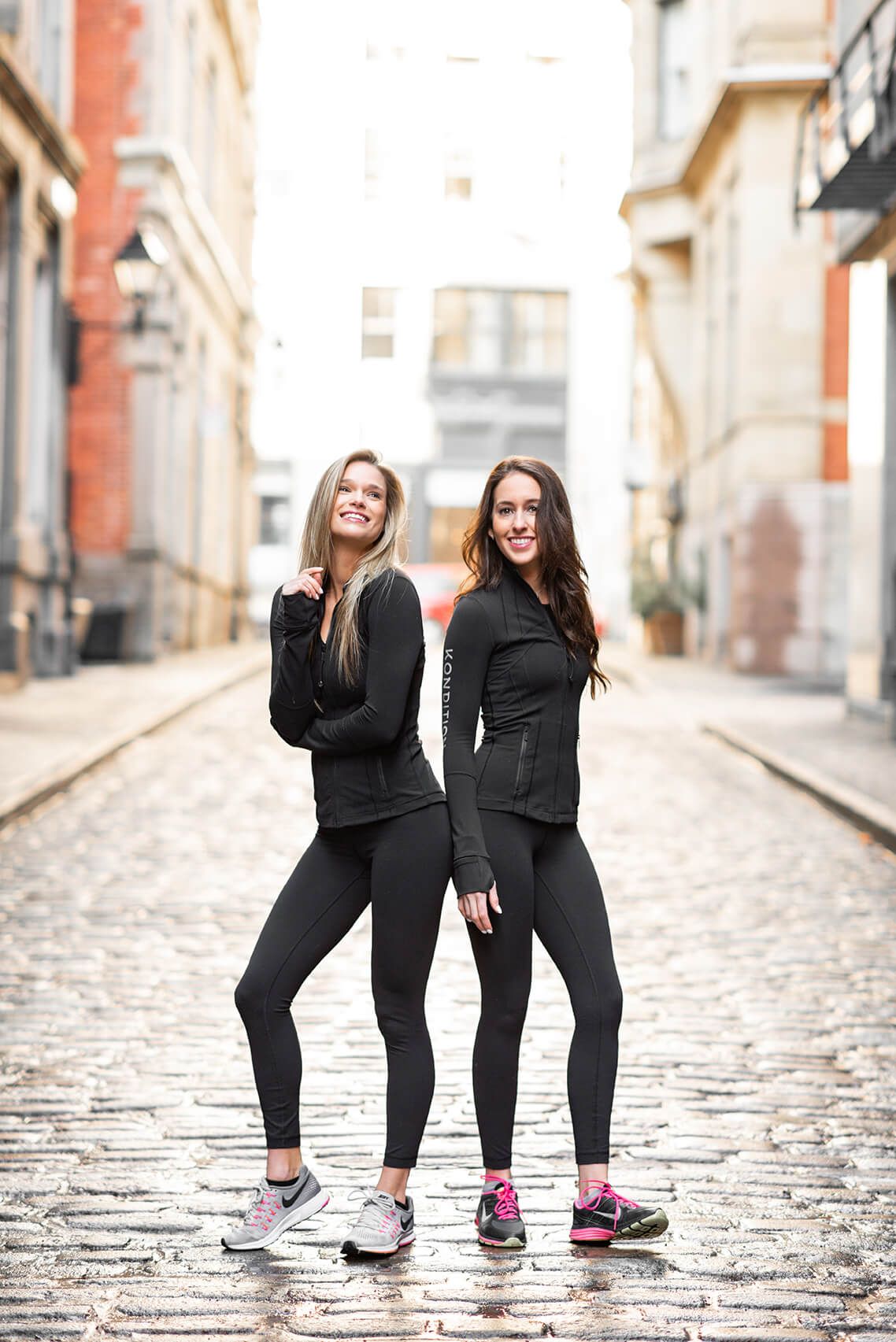 Two women in matching black activewear posing on cobblestone street.