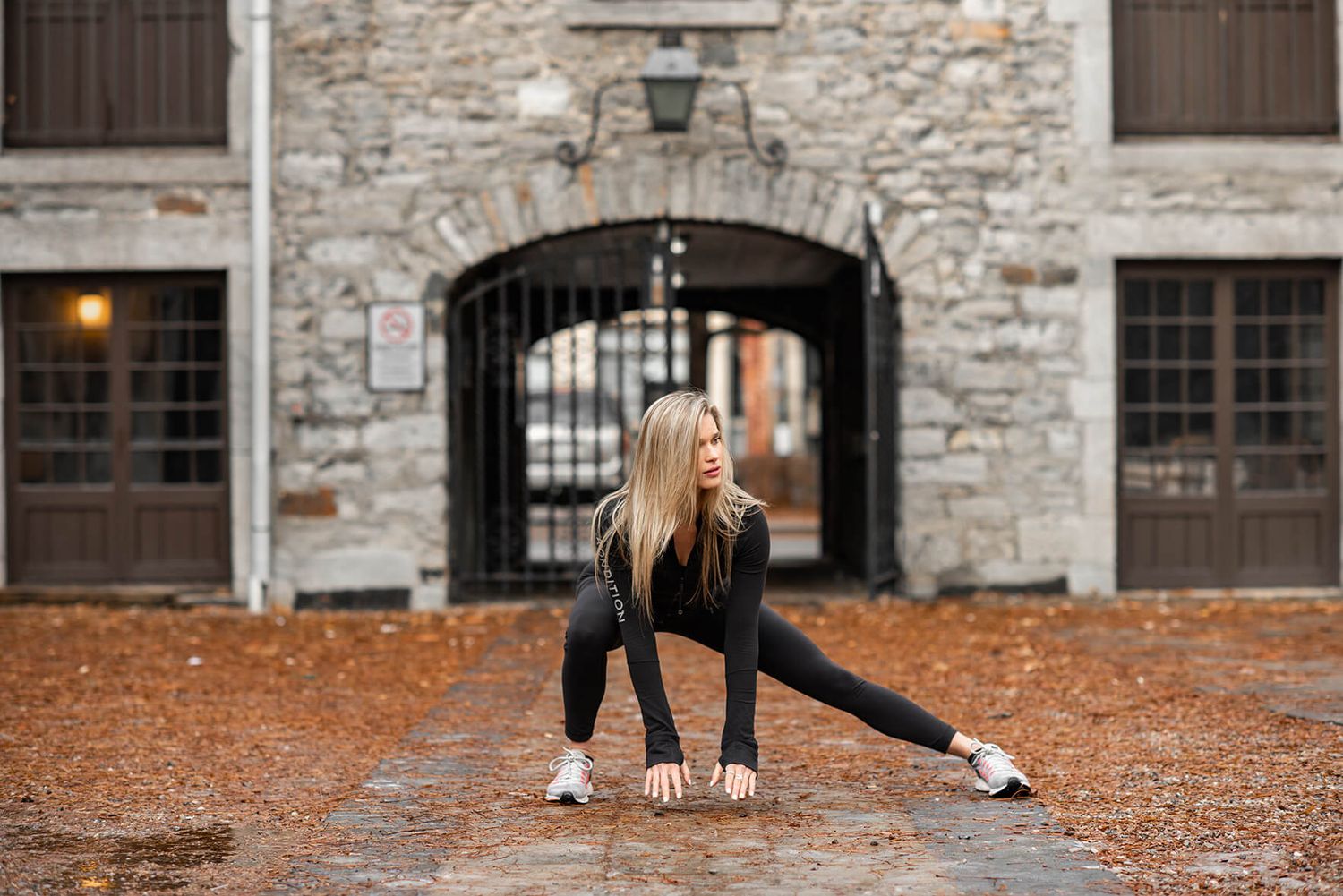 Athletic woman stretches and exercises outdoors against stone building wall on autumn day.