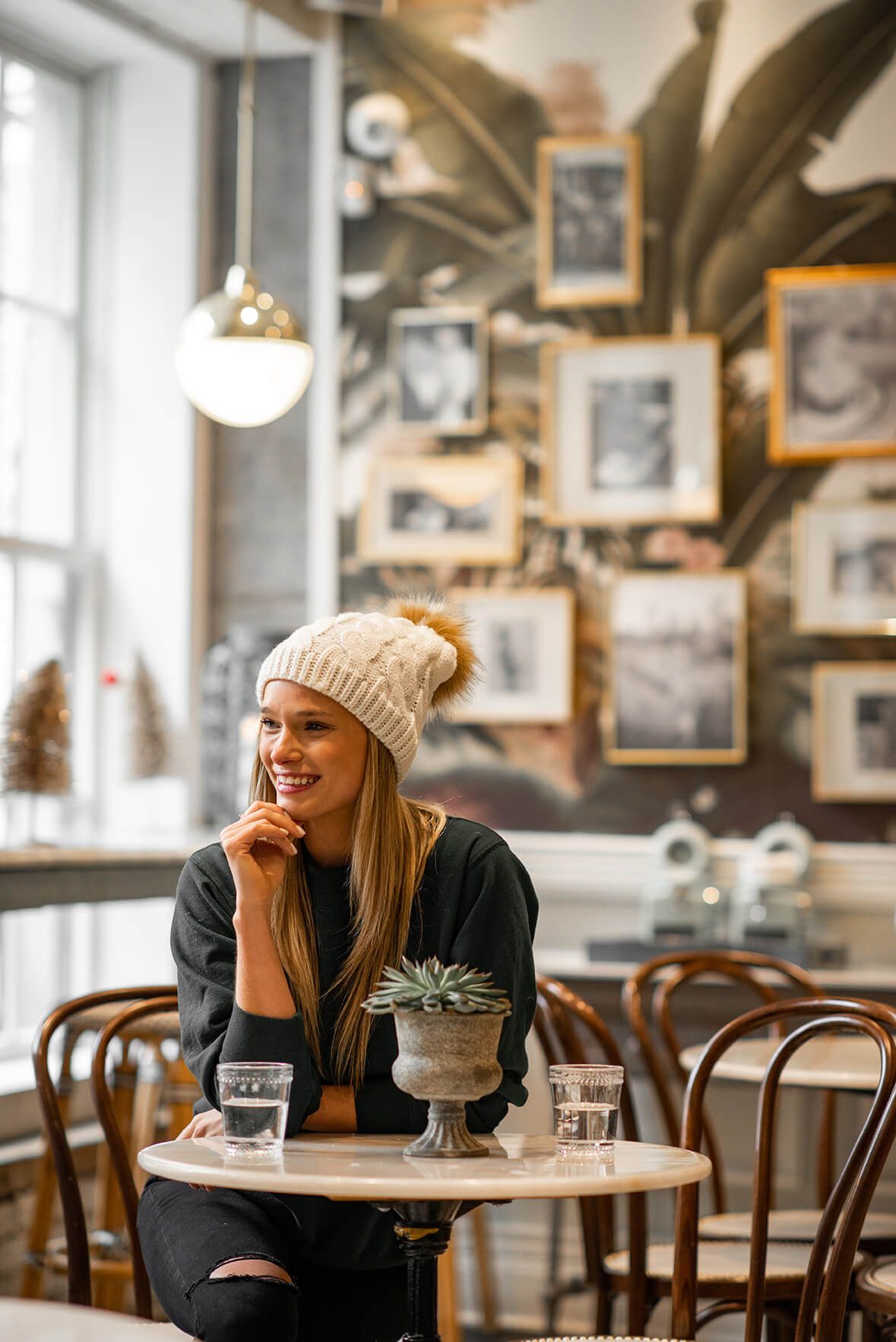 Woman in cream beanie smiles while sitting at marble cafe table with succulent centerpiece.