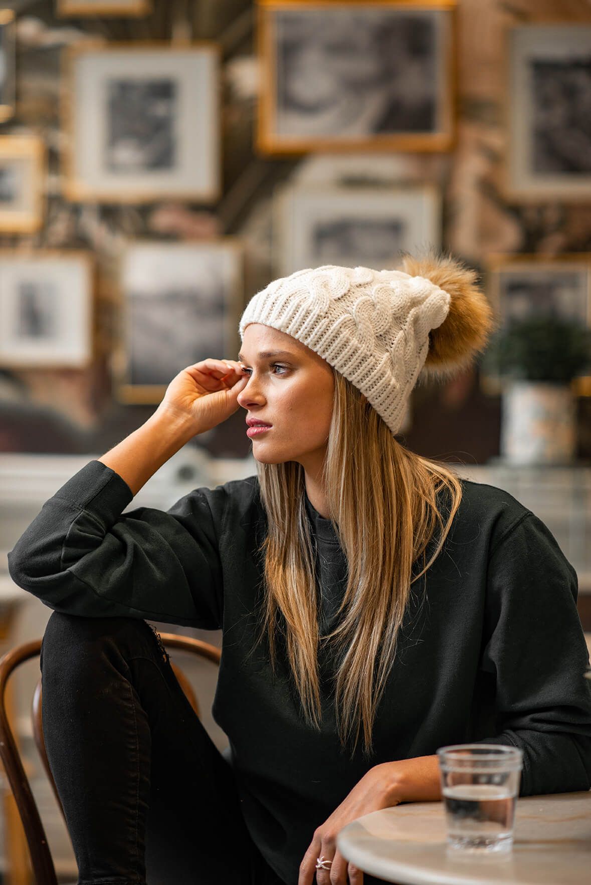 Stylish woman in knit beanie and black sweater poses thoughtfully in vintage cafe setting.