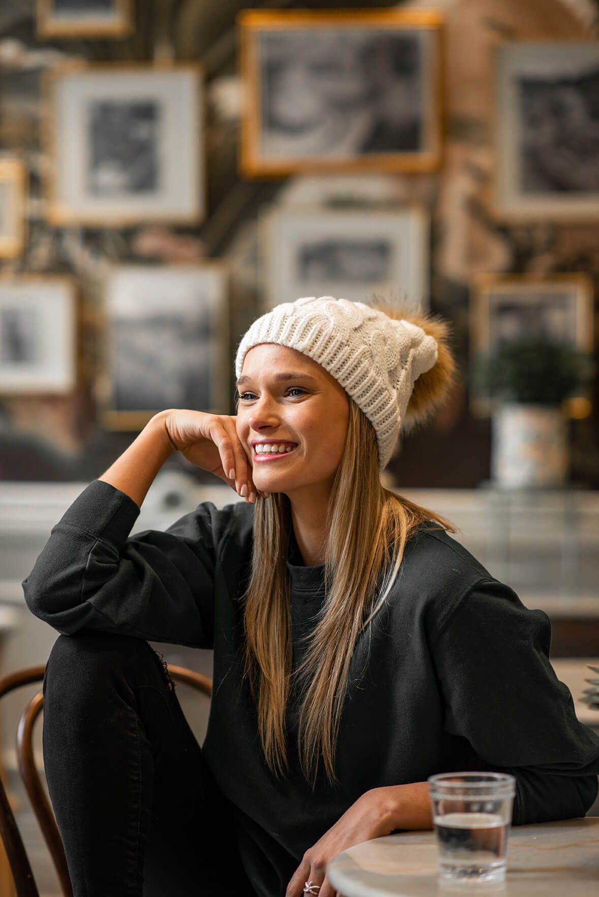 Person wearing beanie and black coat smiles while relaxing in cafe with framed photos on wall.