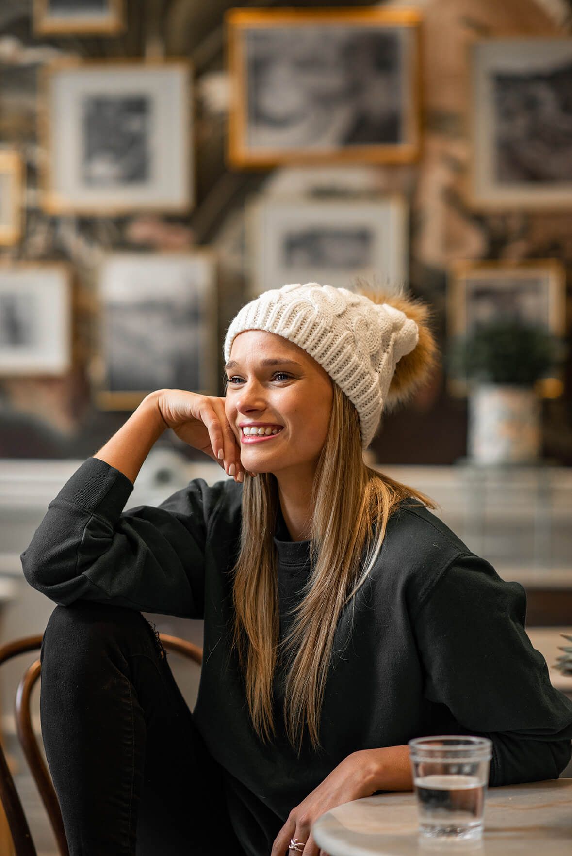Person in black coat and beige beanie smiling while relaxing in cafe setting.