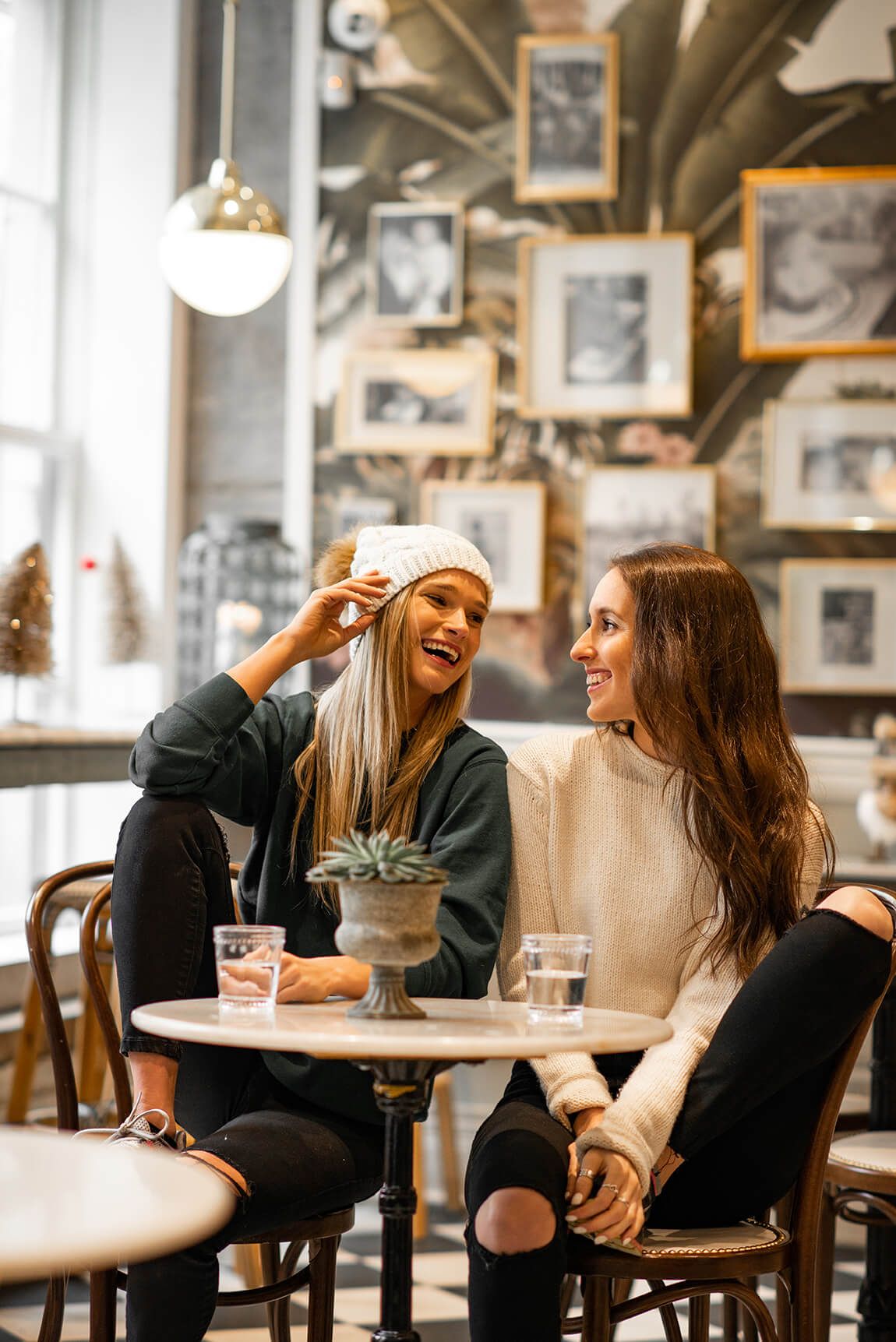 Two friends sharing a laugh at a cozy cafe with vintage photos on the wall and a small succulent plant on their table.