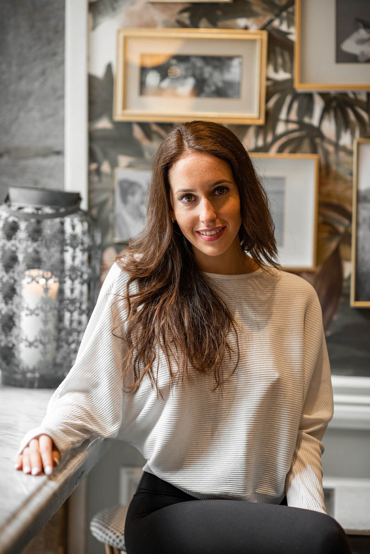 Woman in cream sweater poses casually by window with natural light streaming in.