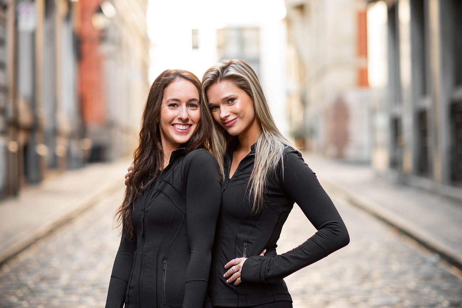 Two friends in black outfits share a laugh while standing on cobblestone street.