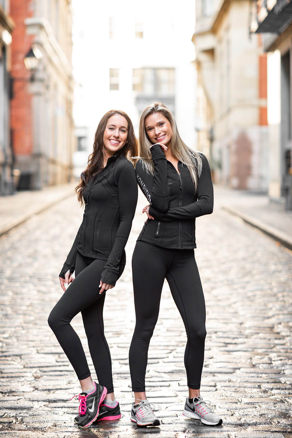 Two friends in matching black athletic wear pose together on a cobblestone street.