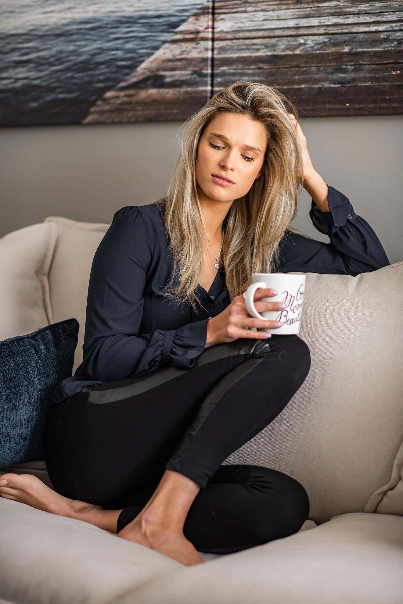 Person relaxing with coffee on grey couch beneath coastal artwork.