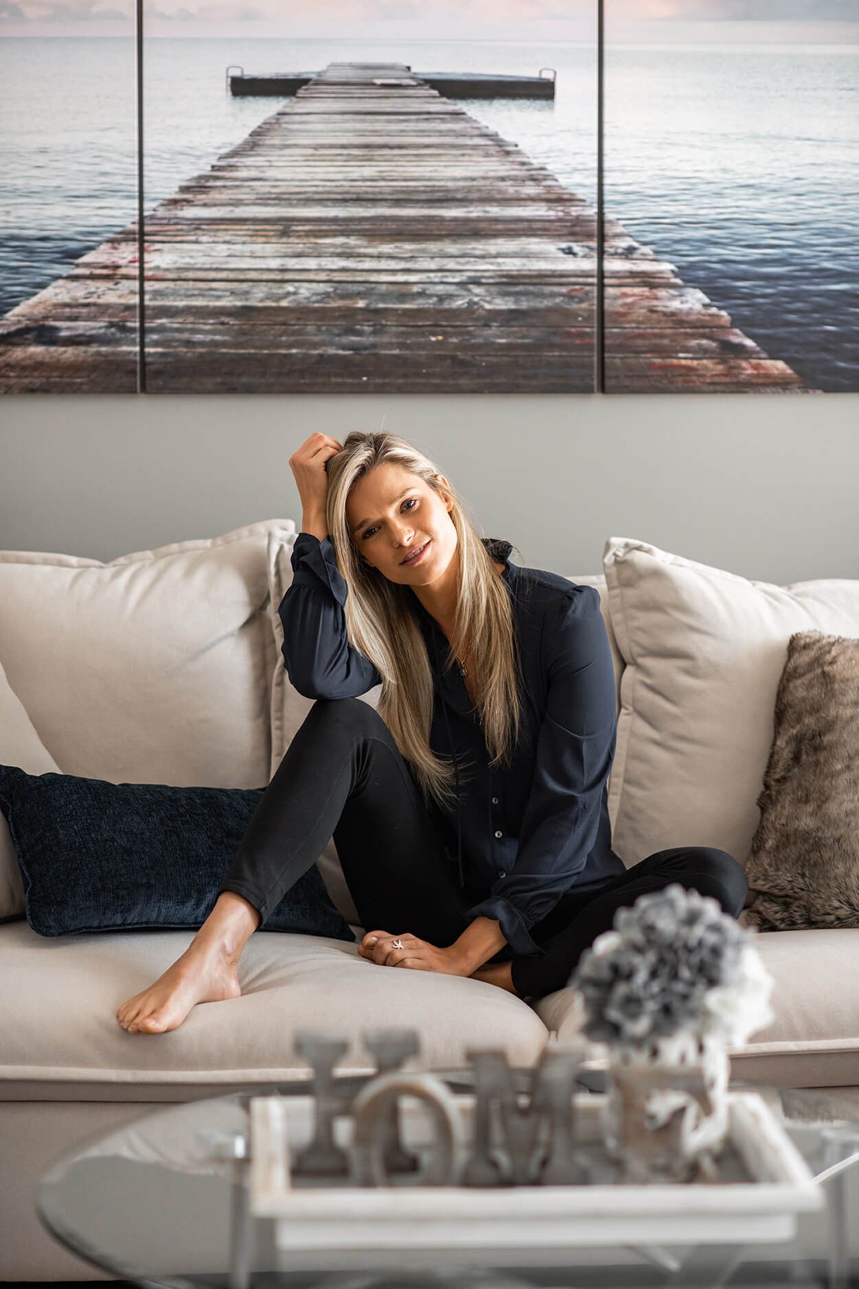 Person relaxing on white couch beneath coastal pier artwork in modern living room setting.