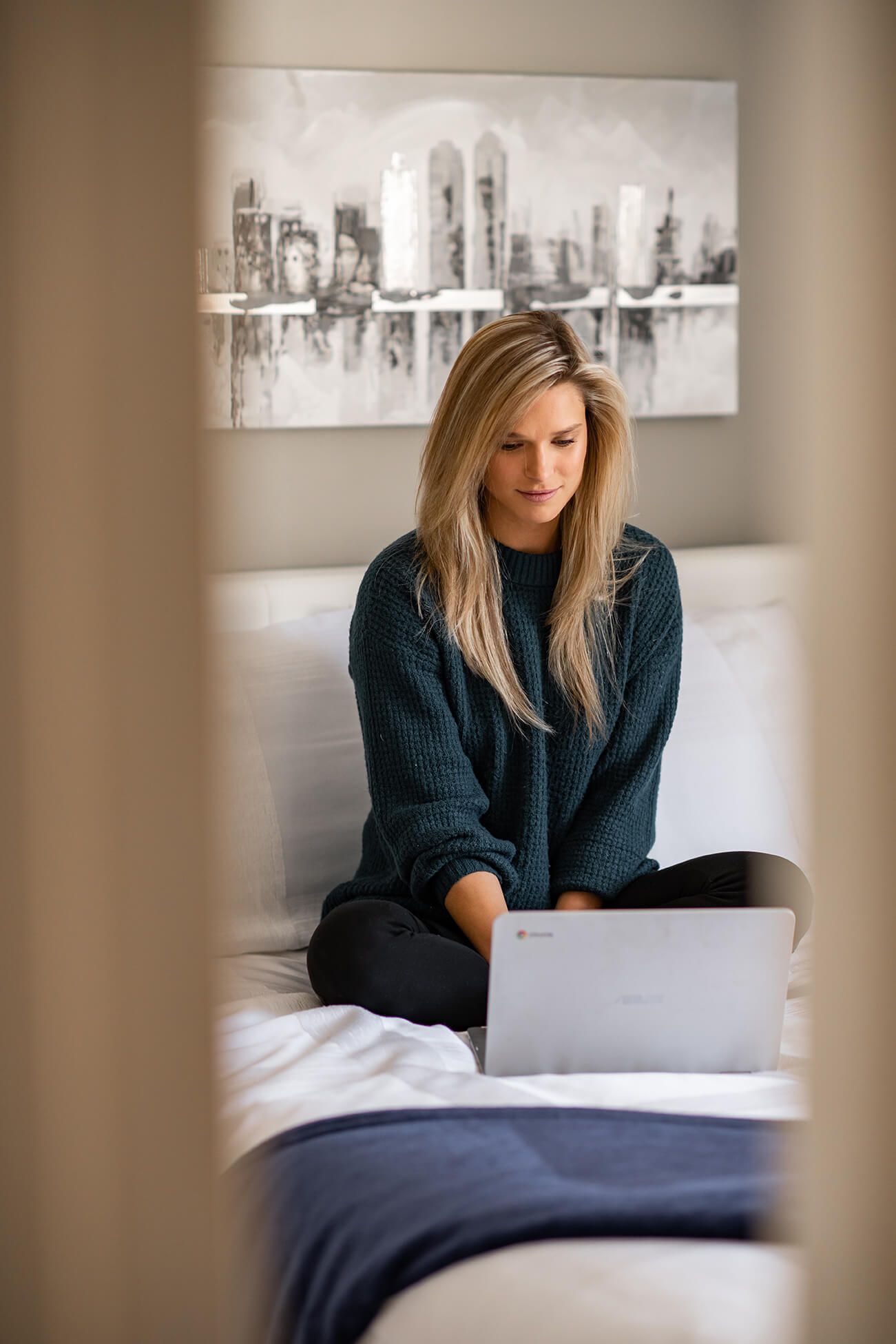 Person in navy sweater working on laptop while sitting on white bed beneath urban skyline artwork.