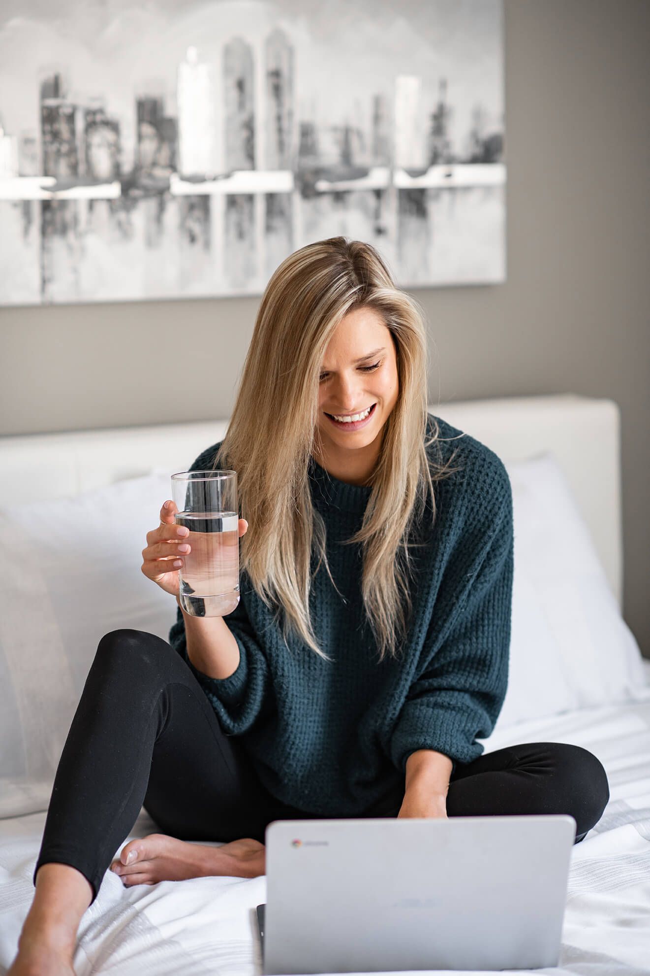 Person relaxing with laptop and drink on cozy bed with city artwork in background.