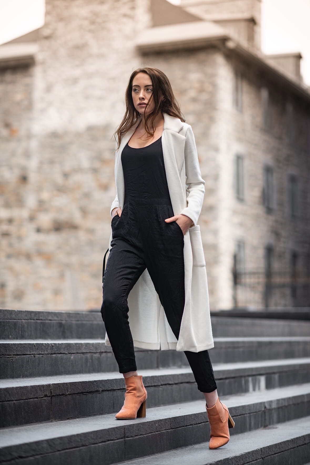 Modern street style look with black jumpsuit, white blazer and tan boots on concrete steps.