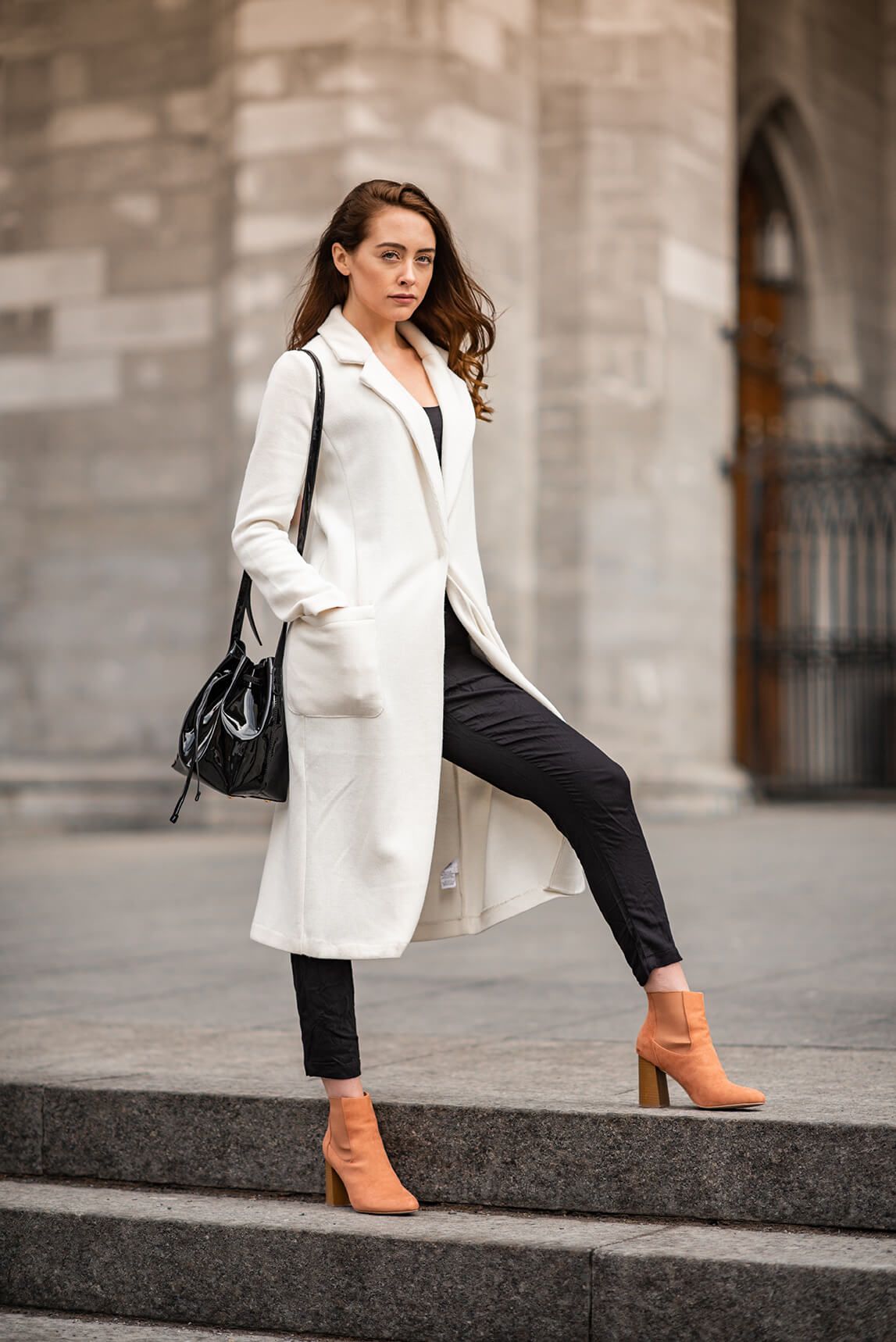 Fashion model in long white coat and orange boots poses on urban steps.