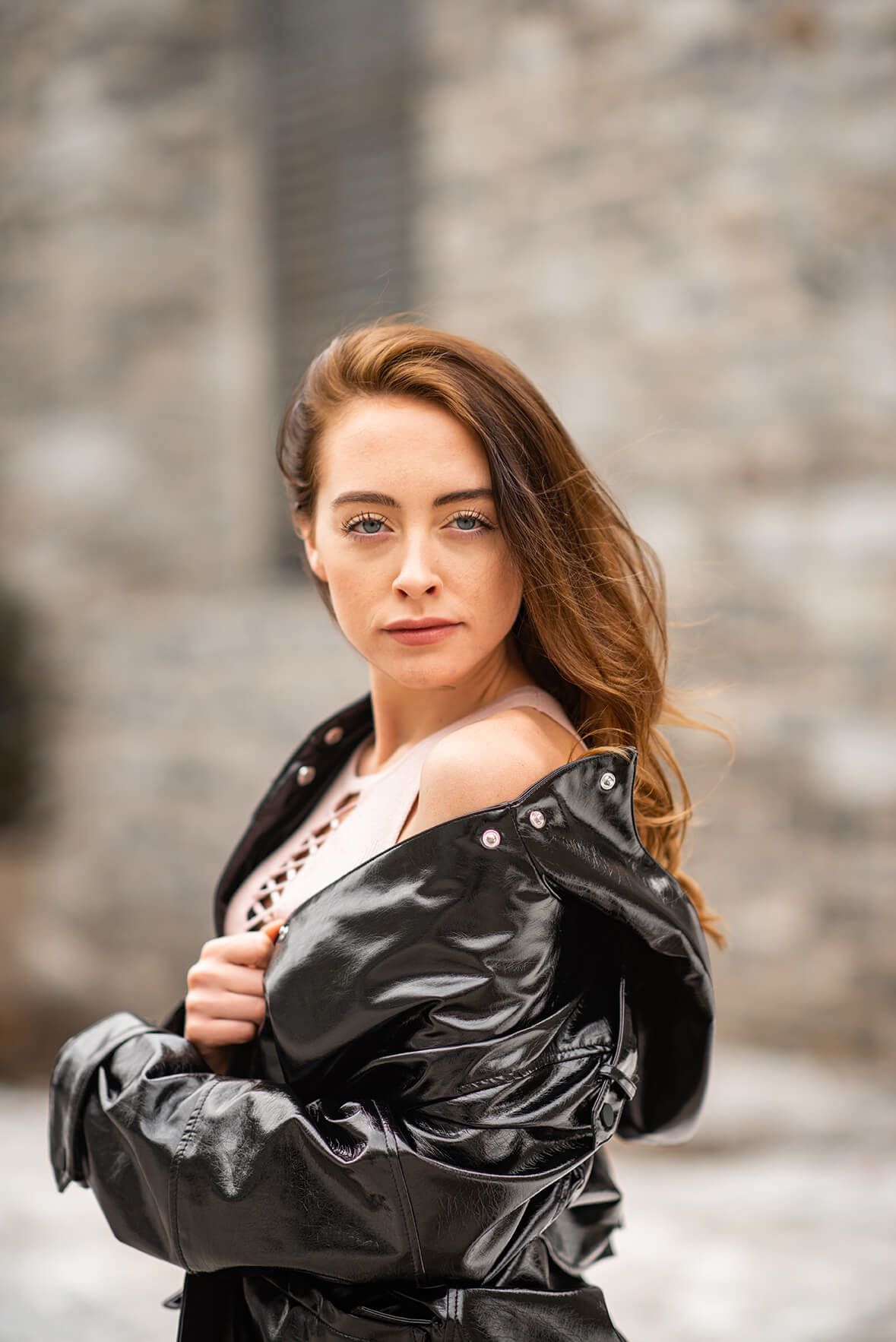 Fashion model wearing black leather jacket poses against stone wall with dramatic lighting.