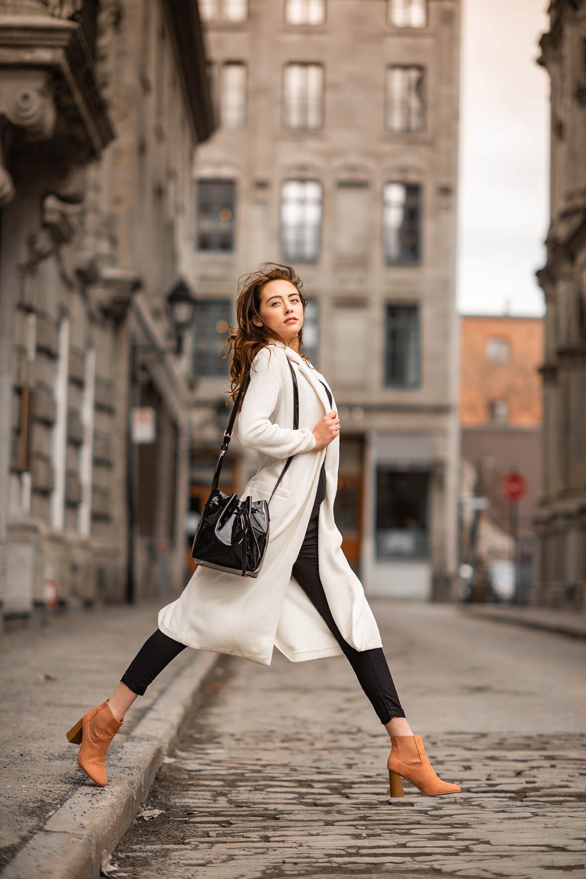 Fashion model in cream coat and brown boots posing on cobblestone street.