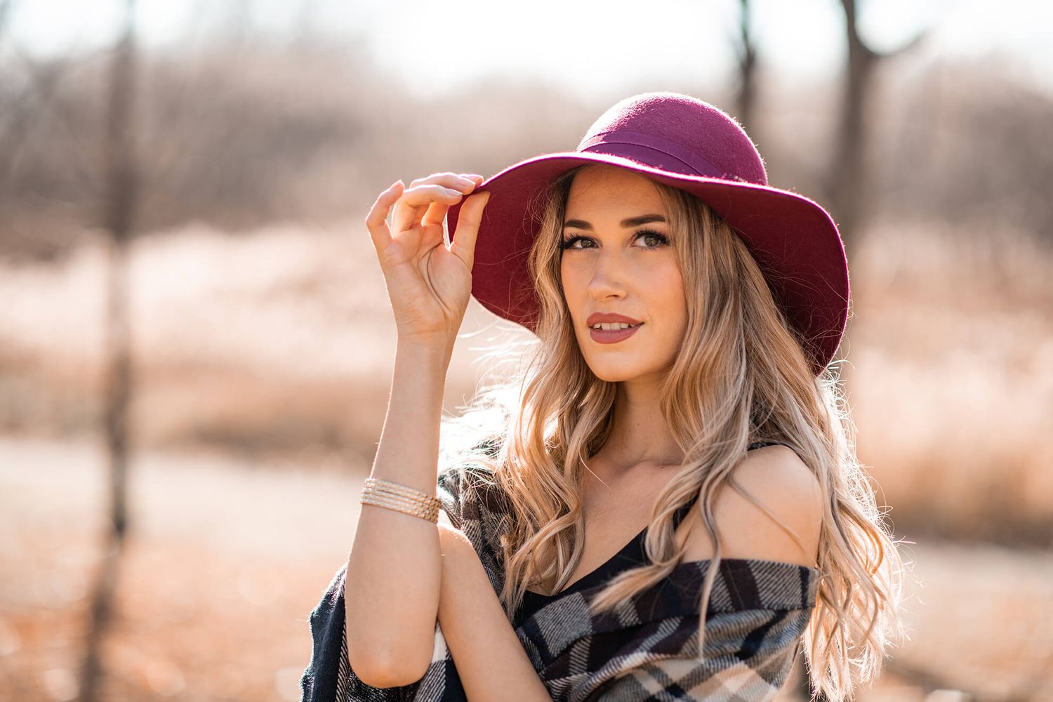 A person in a burgundy hat and plaid wrap stands against a natural autumn backdrop.