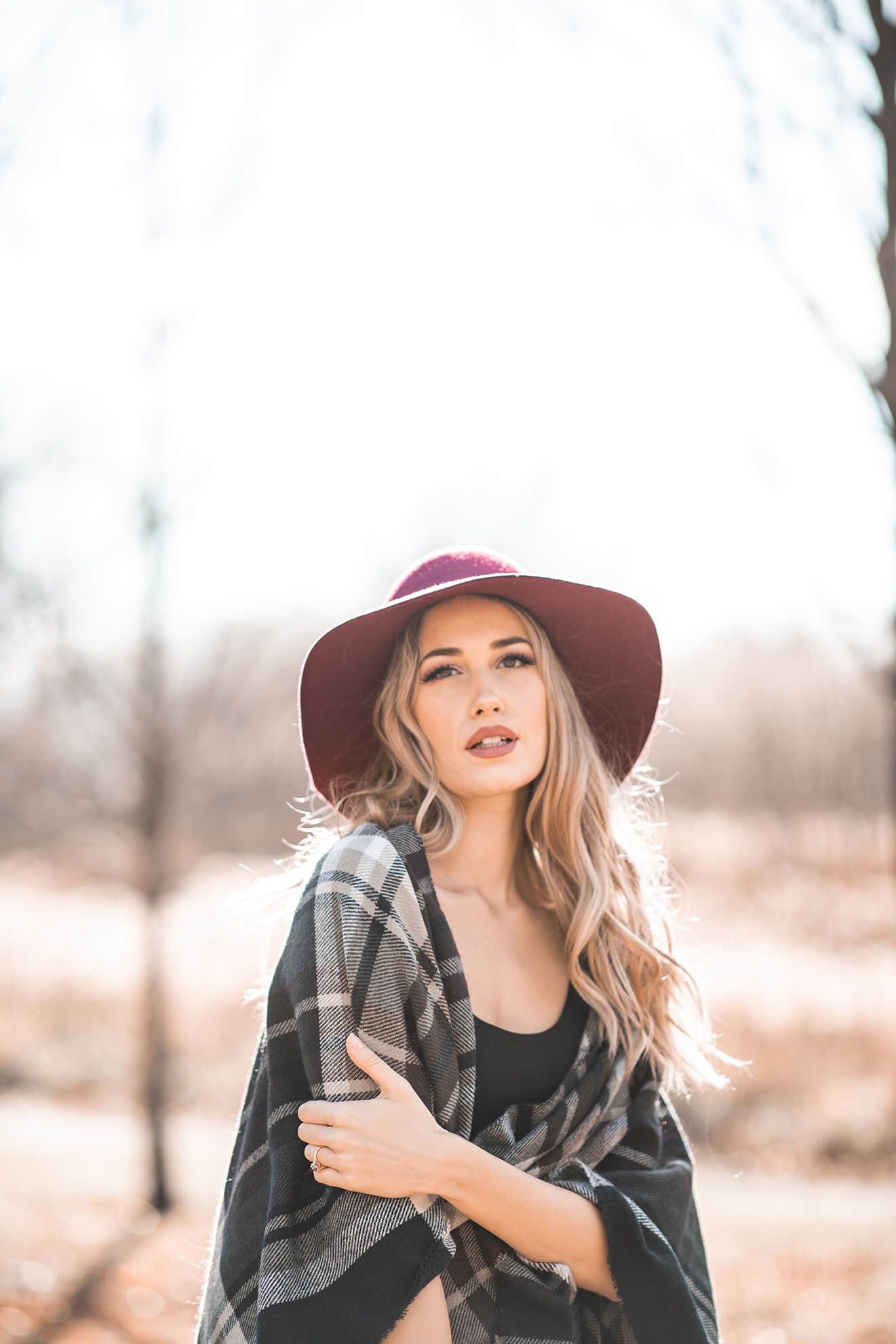 A person in a burgundy wide-brim hat and plaid scarf poses outdoors in autumn.