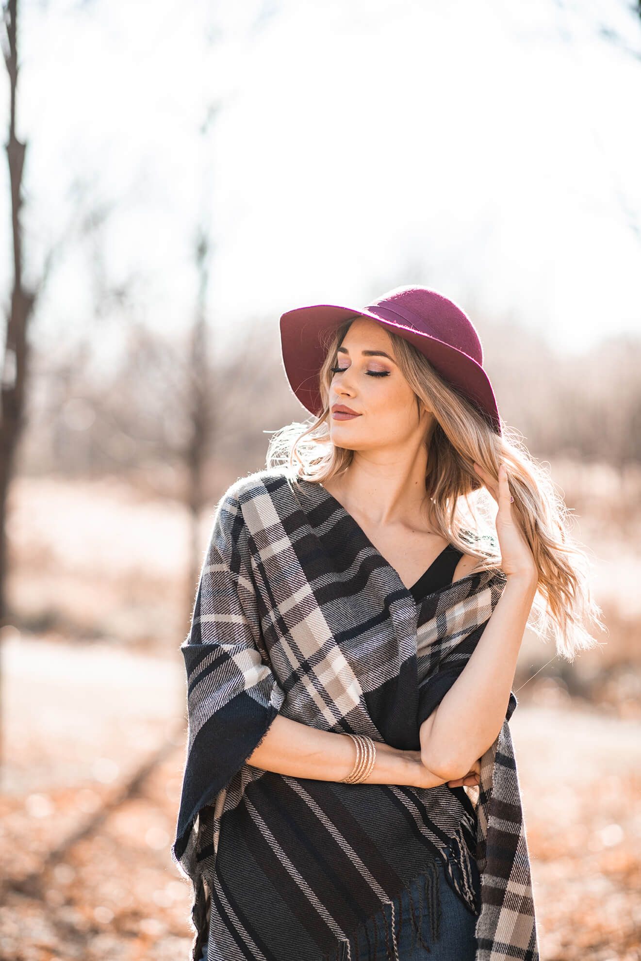 Autumn fashion shoot with burgundy wide-brim hat and plaid wrap against bare trees.