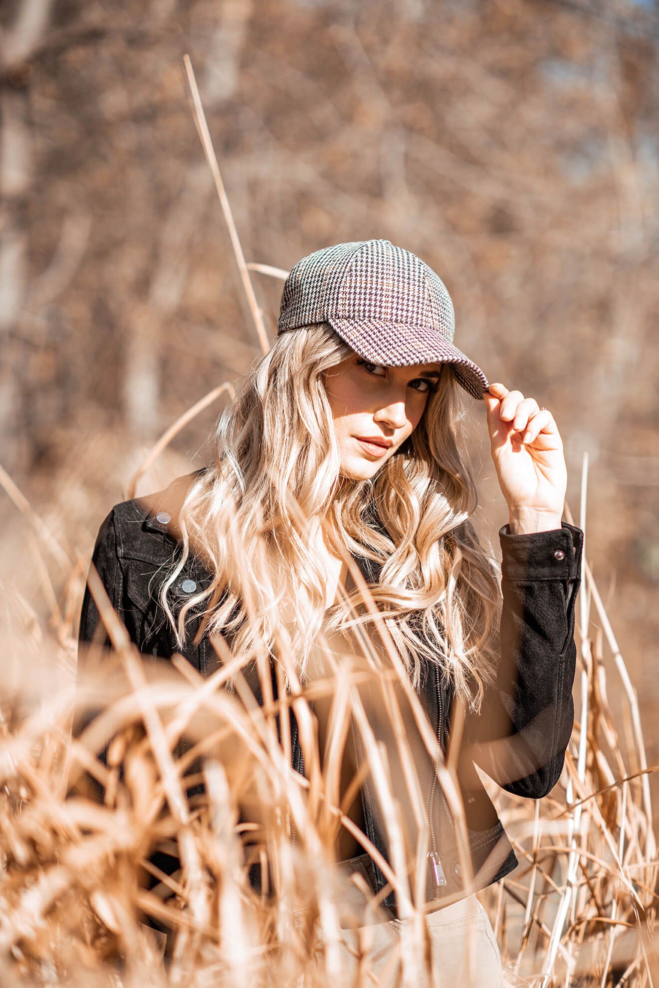 Artistic portrait through dried grass featuring a houndstooth cap and natural lighting.