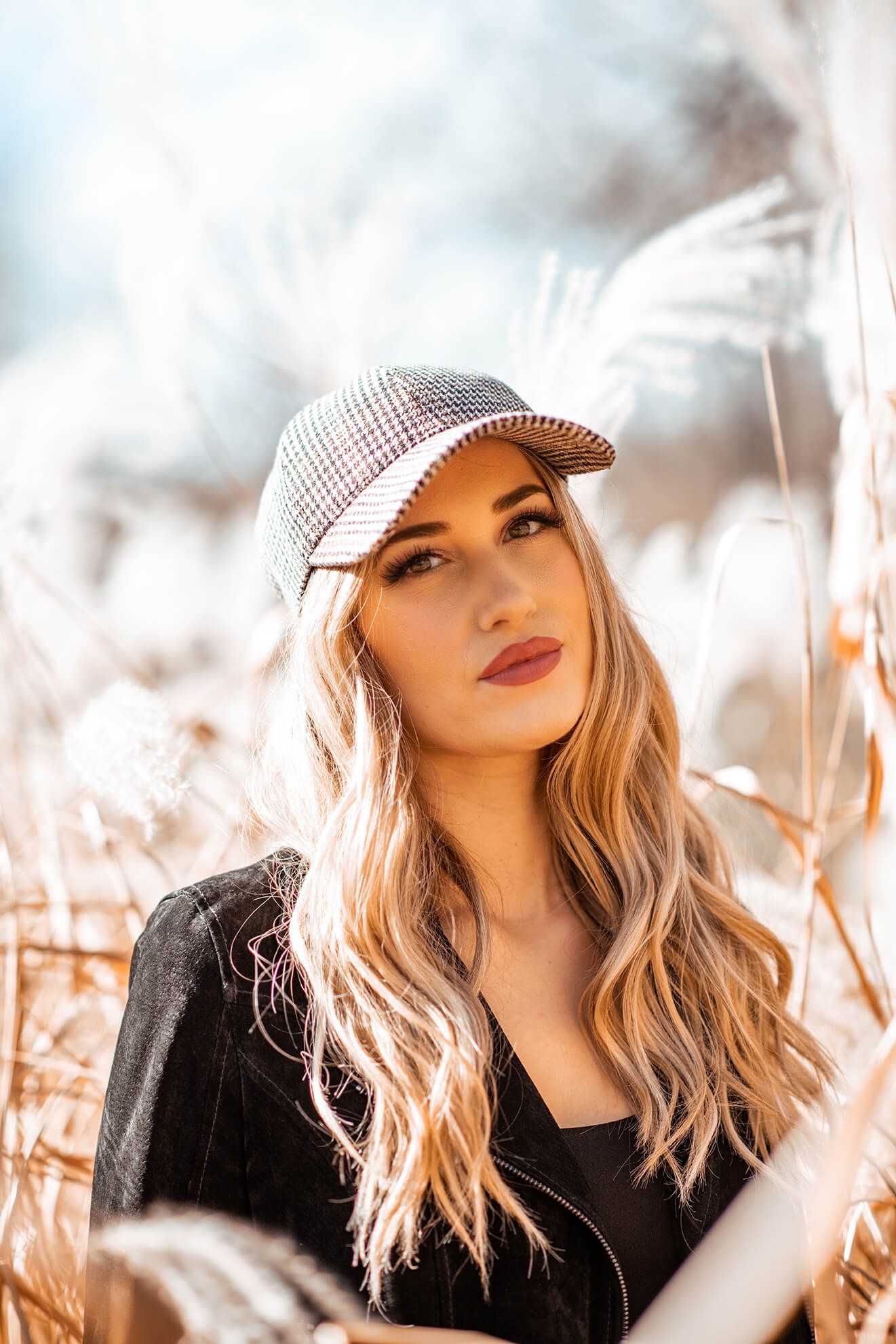 A fashion portrait in tall grass with warm autumn tones featuring a plaid newsboy cap and blonde hair.