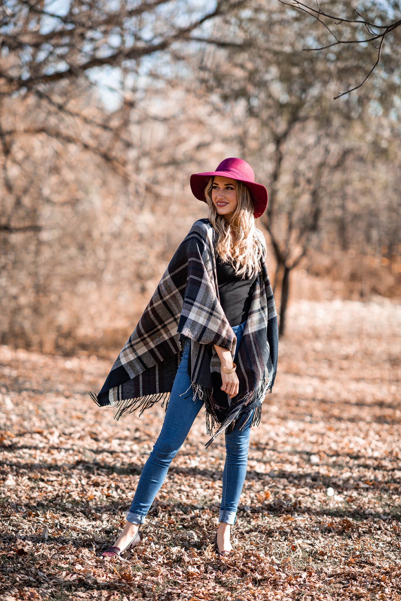 Woman in burgundy hat and plaid jacket poses creatively on autumn ground.