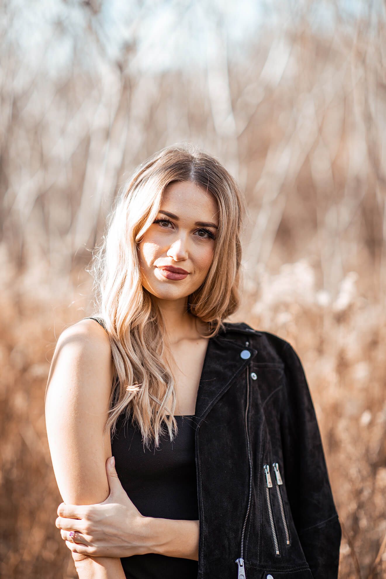 Woman in black leather jacket poses casually against natural autumn background.