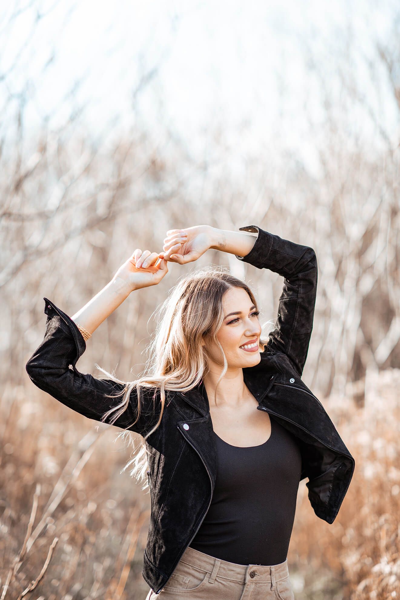 A woman in black clothing dances freely in a natural setting with bare trees.