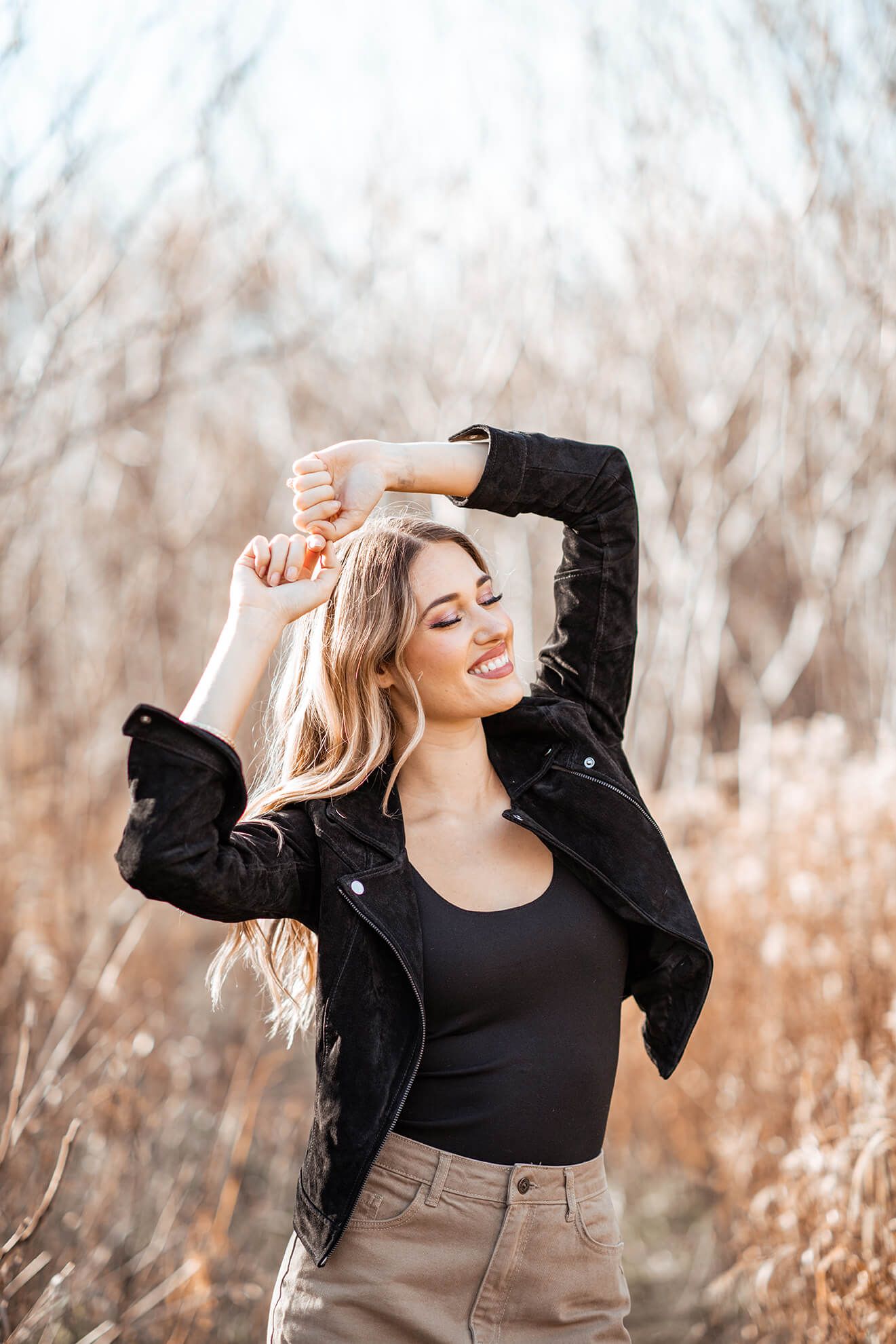 A woman in a black jacket dances joyfully among bare trees in a natural setting.
