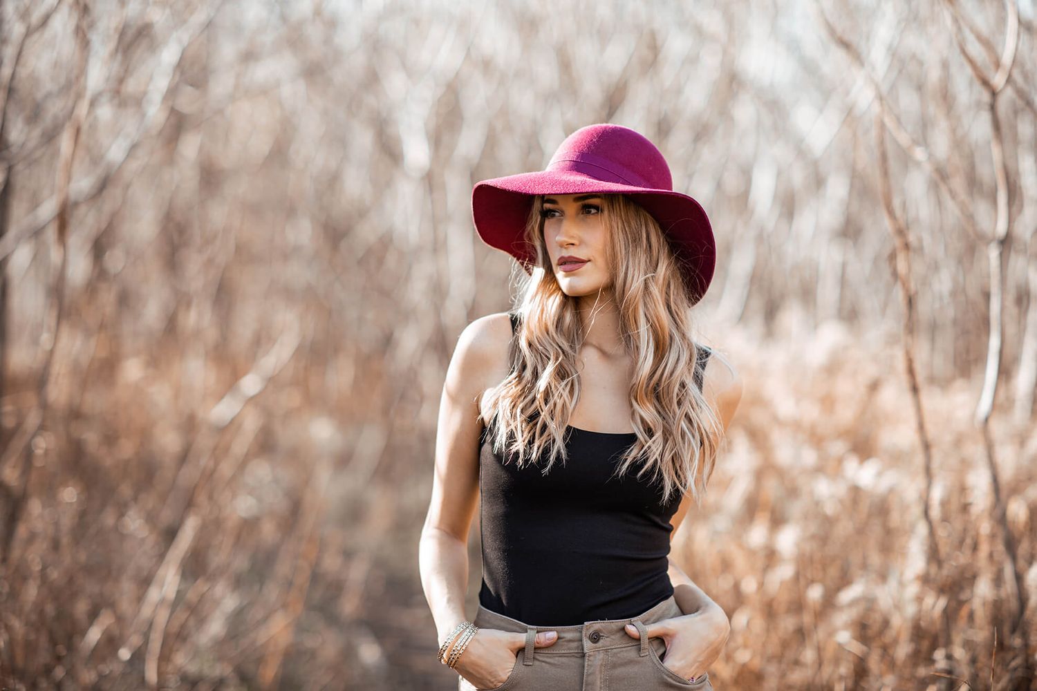 A stylish woman in a burgundy hat poses against a natural autumn backdrop with bare trees.