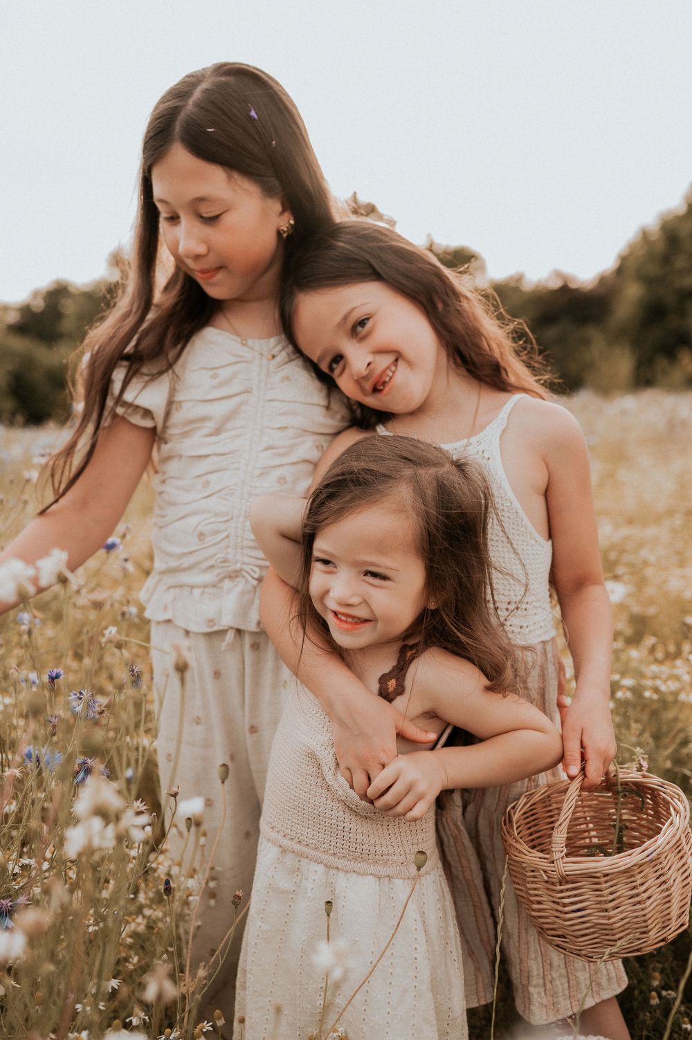 Hartverwarmend moment in een veld vol wilde bloemen bij zonsondergang, met lichte crèmekleurige jurken — gezinsshoot in West-Vlaanderen