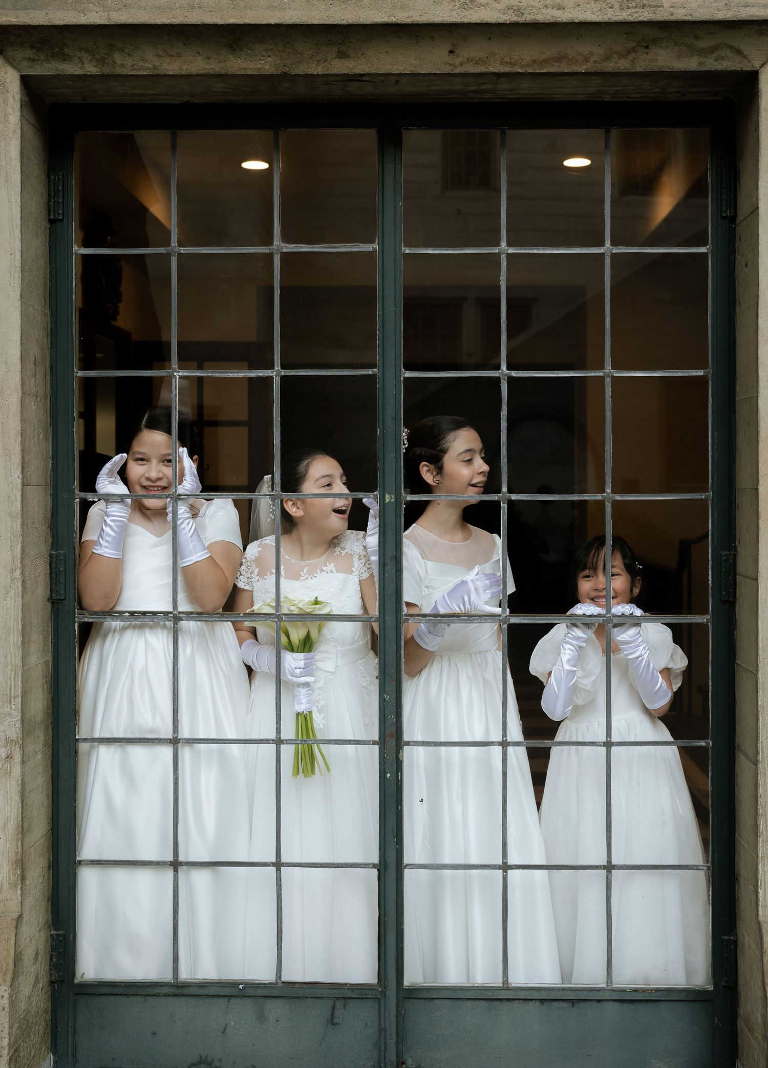 A row of bridesmaids in white dresses peek through windows of a rustic building, creating a playful wedding photo.