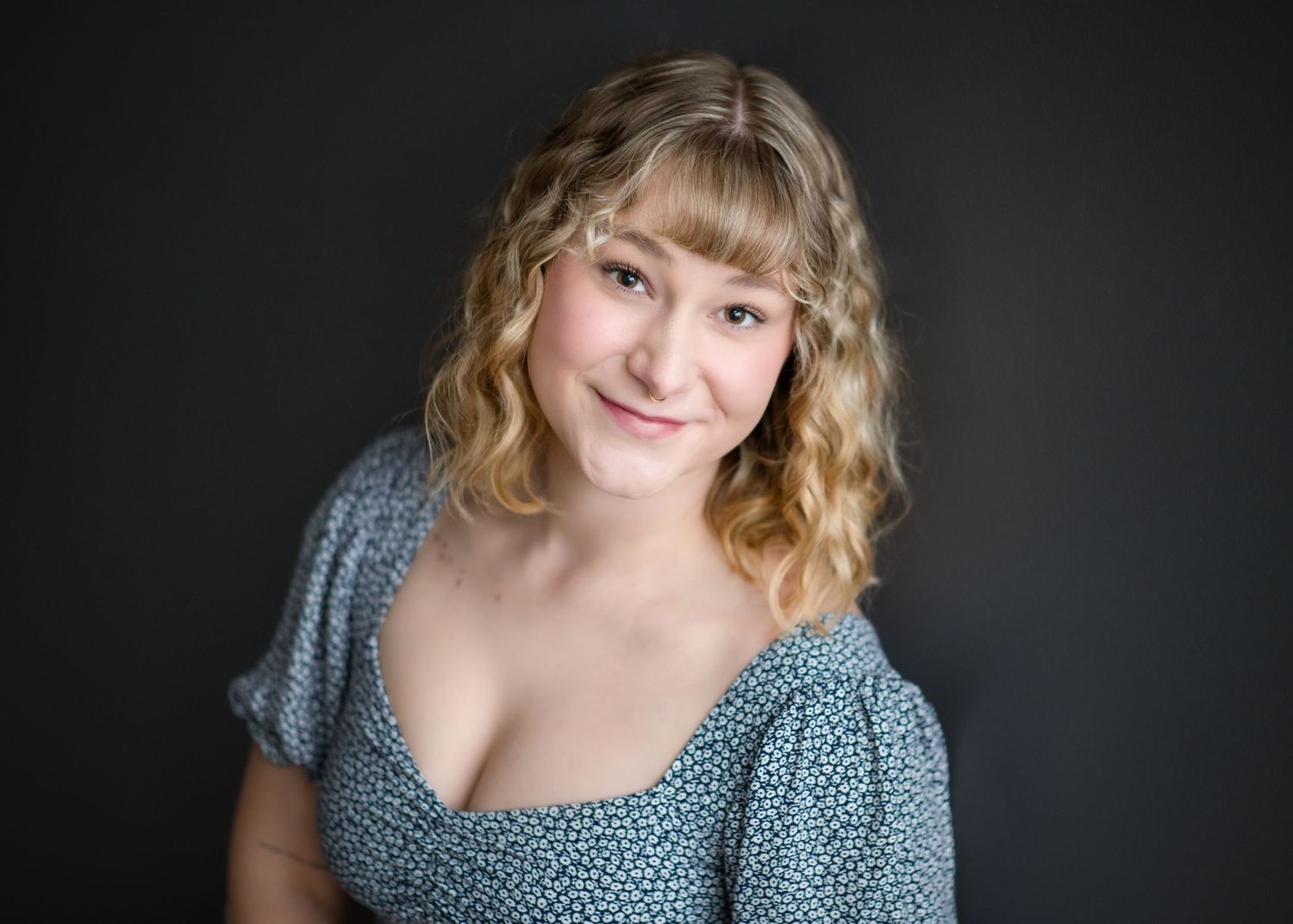 Professional headshot of someone with blonde wavy hair wearing a blue floral dress against a dark background.