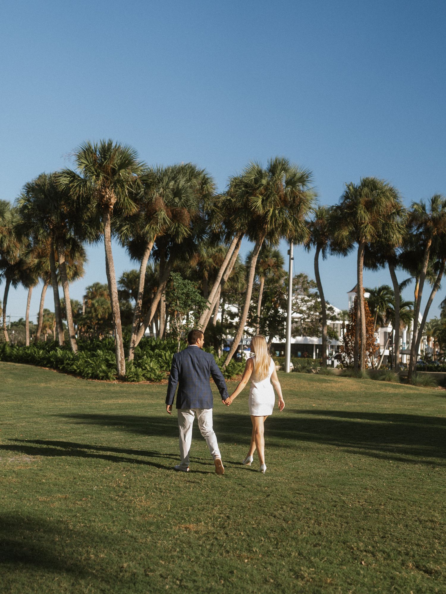A couple dances playfully on a grassy lawn lined with palm trees under a bright blue sky.