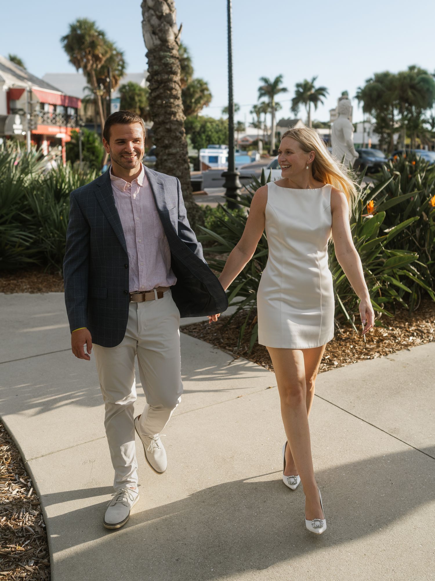 A couple walks together on a palm tree-lined sidewalk in casual summer attire on a sunny day.