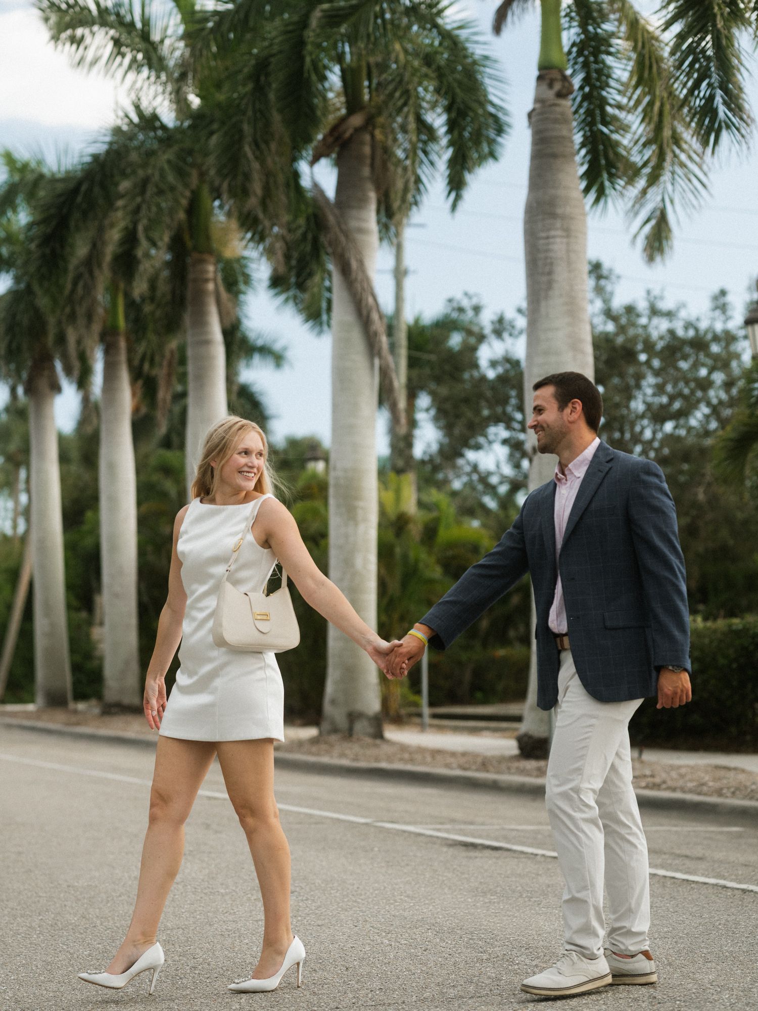 A couple in formal attire playfully interacts along a palm tree-lined pathway during a photo session.